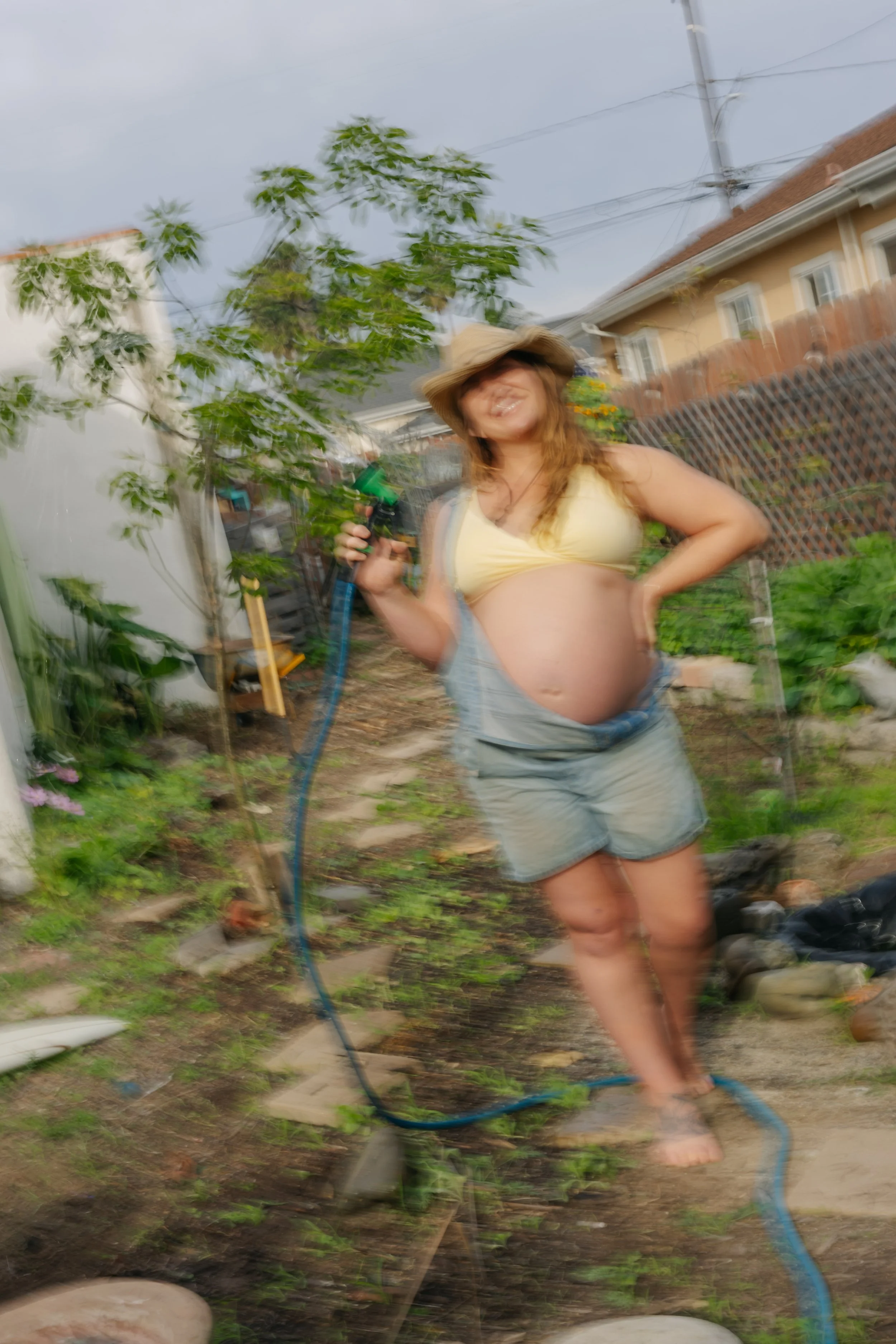 A woman with curly hair wearing a sunhat, yellow tank top, and shorts, standing in a garden holding a garden hose and smiling.