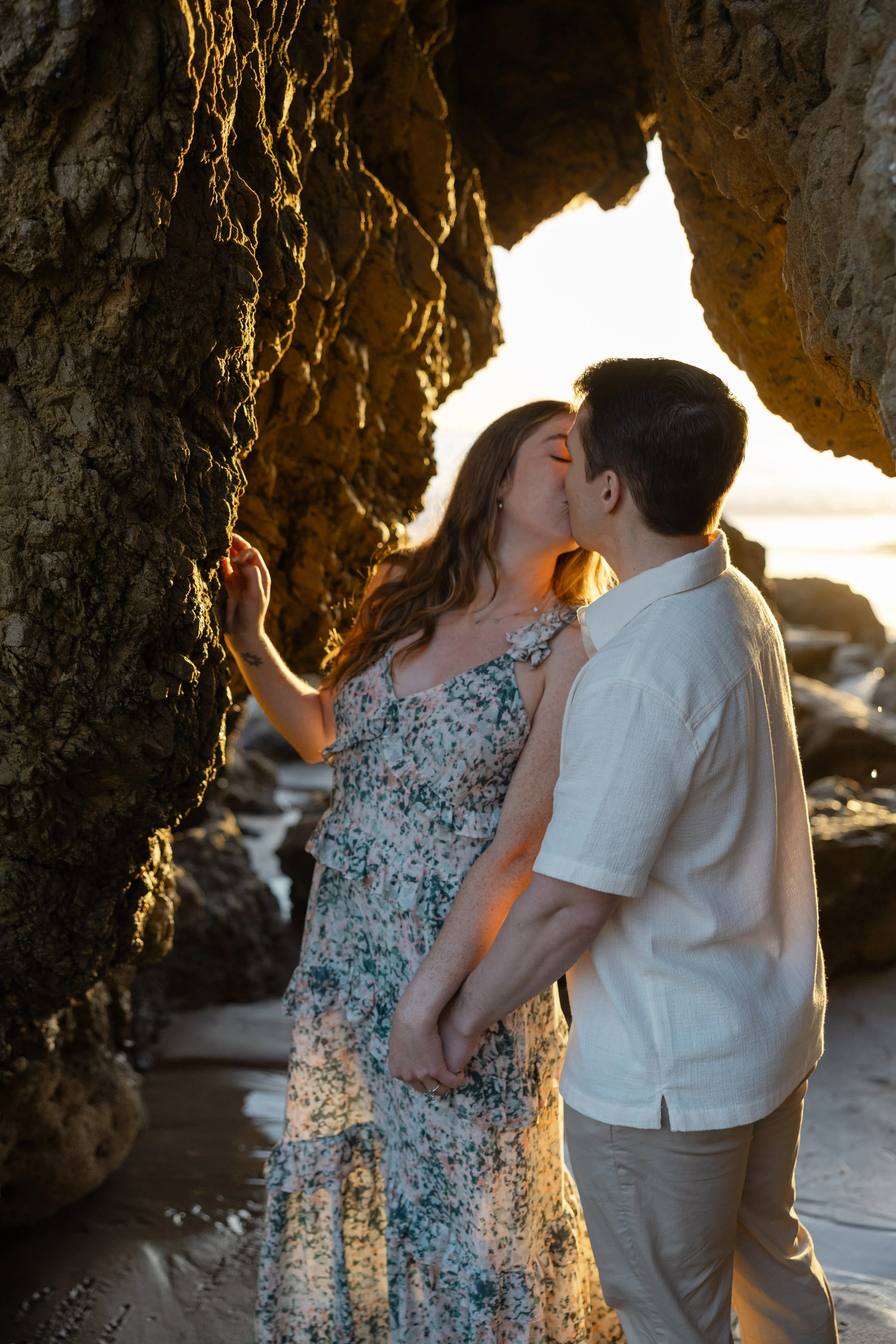 A couple kissing on a beach at sunset, standing between large rock formations.