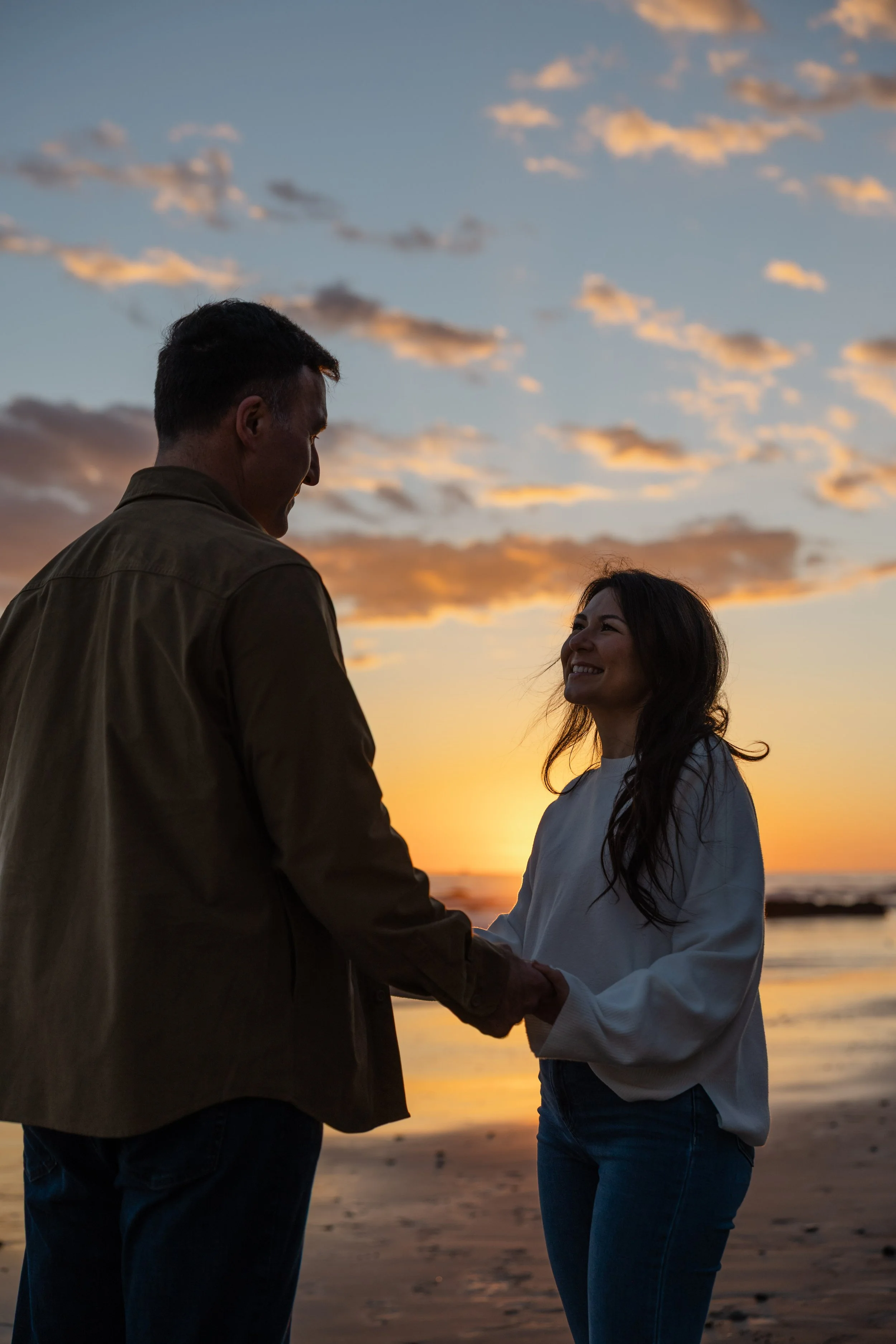 A couple holding hands at the beach during sunset, facing each other and smiling.