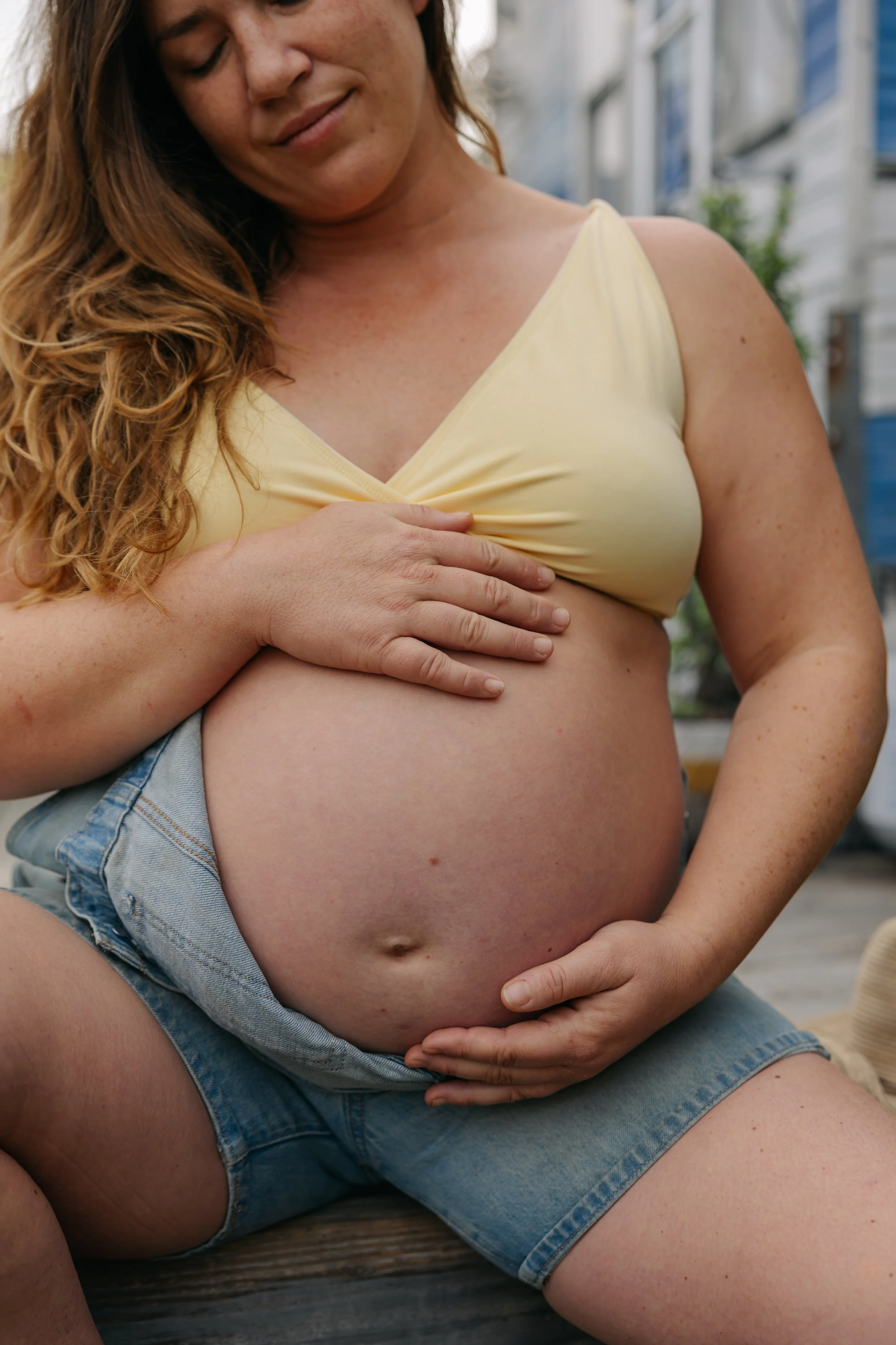A pregnant woman sitting outdoors, touching her belly with both hands, wearing a yellow top and denim shorts, with a gentle smile.