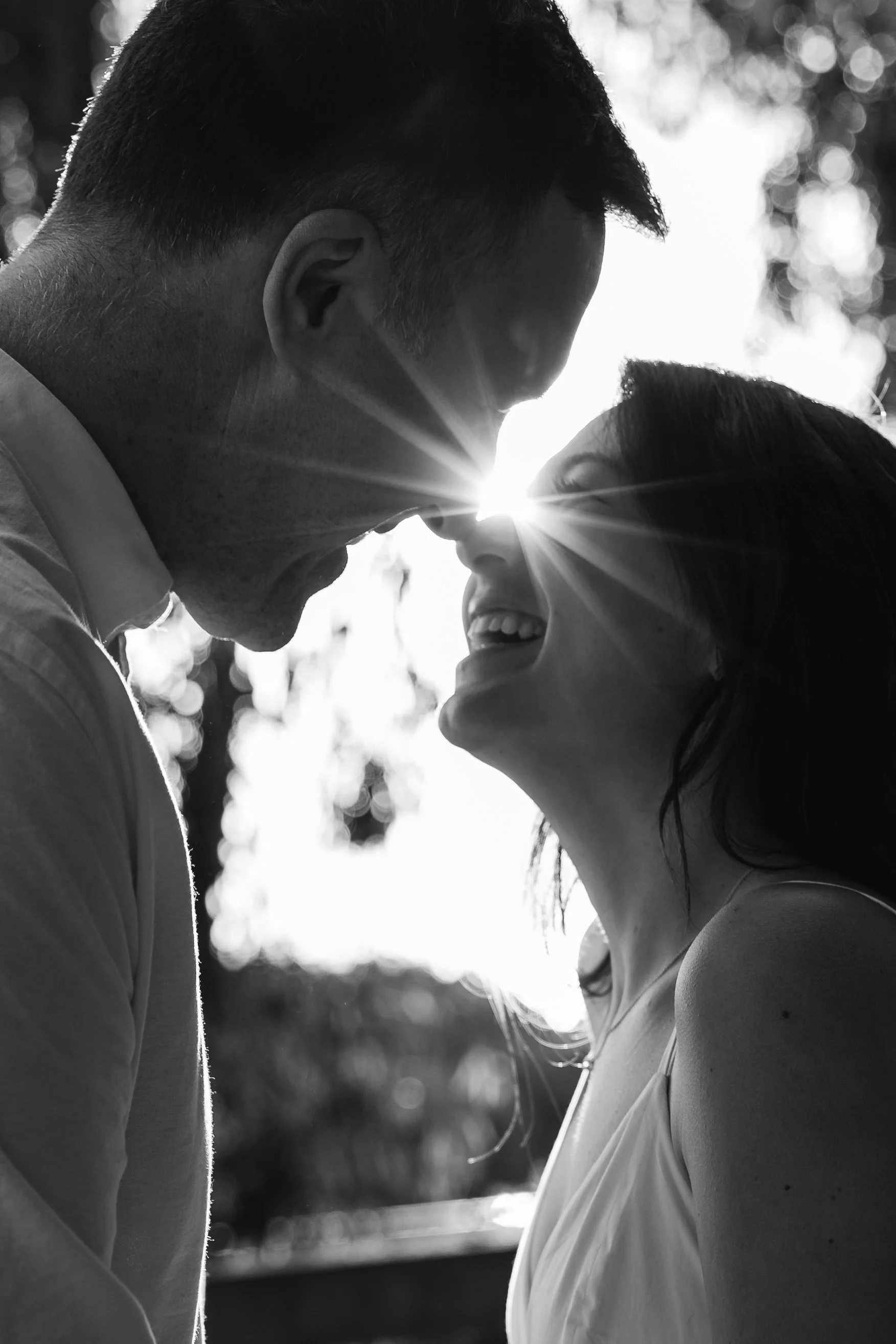 A black and white photo of a couple close together, smiling and laughing, with their foreheads touching and the sun shining behind them creating a starburst effect.
