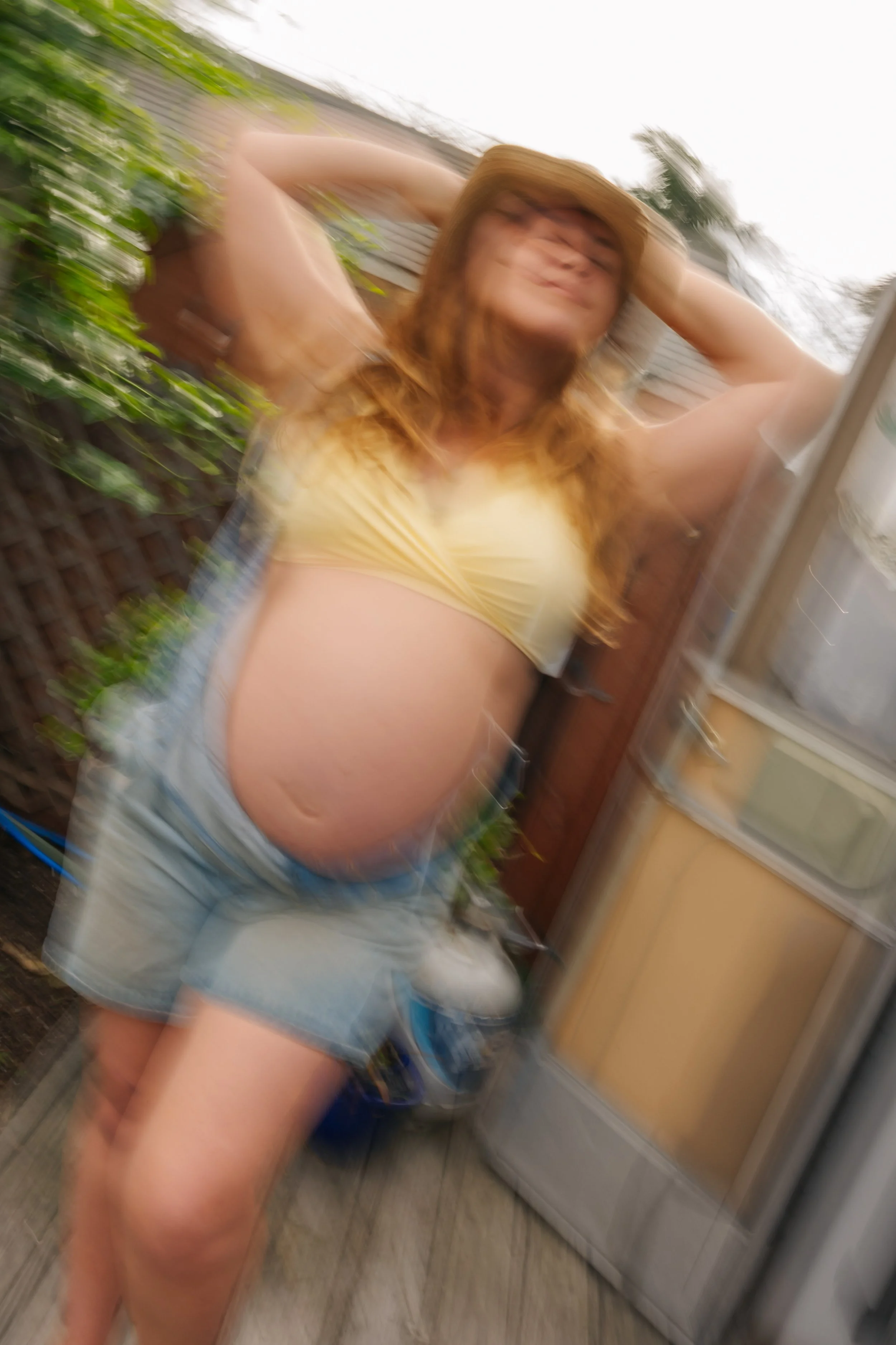A woman with red hair, wearing a yellow crop top and denim shorts, standing with her hands behind her head in an outdoor setting, with a wooden fence, garden plants, and outdoor storage cabinets visible in the background. The image is blurry and appe