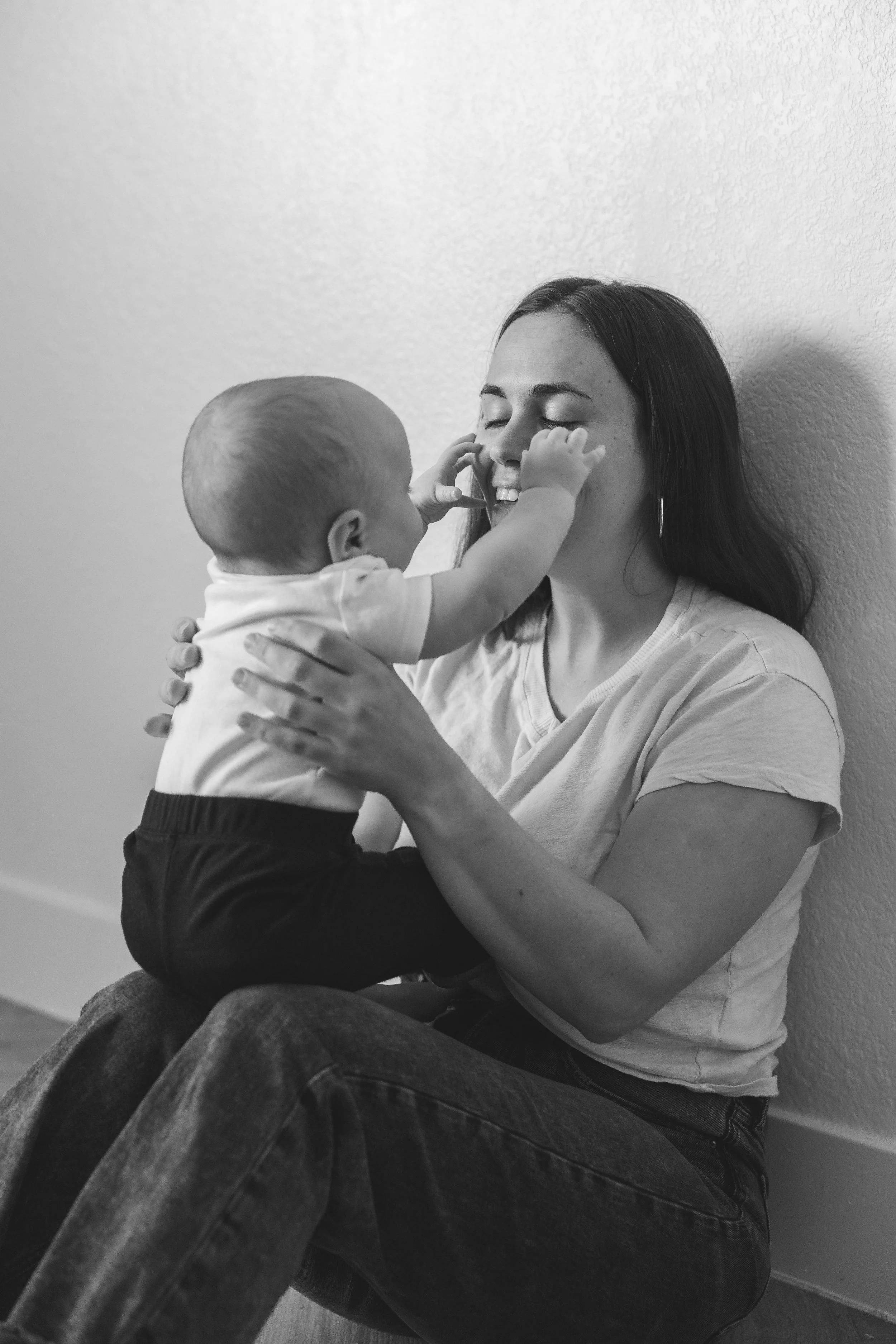 A woman with long dark hair and a white T-shirt sitting on the floor, smiling while holding a baby boy who is touching her face.