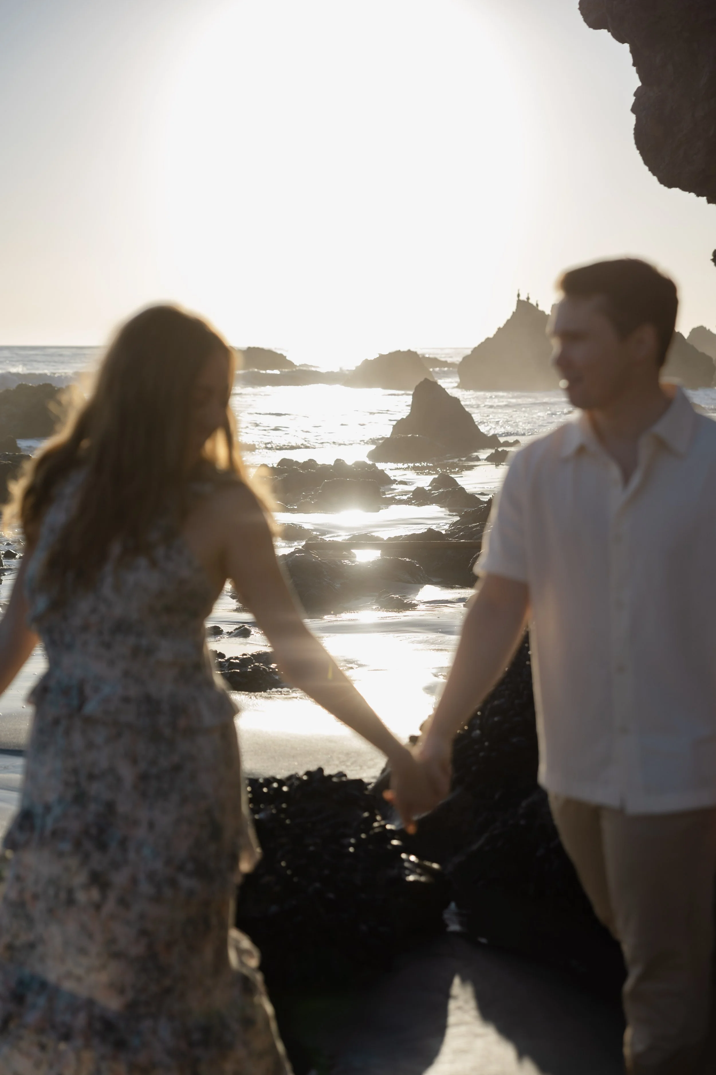 A couple holding hands on a beach at sunset, with rocky formations and the ocean in the background.