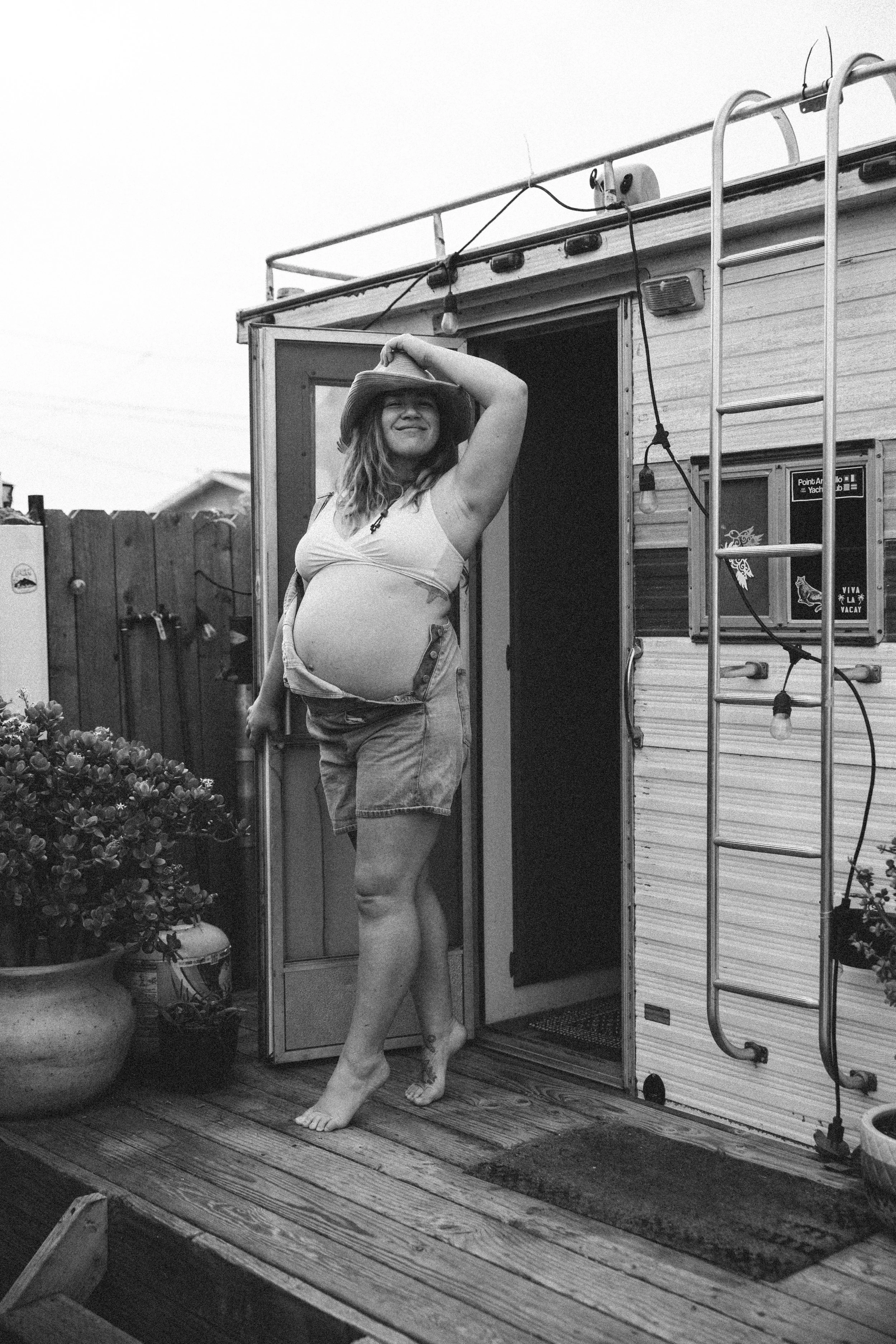 A woman wearing a hat and casual clothes, standing barefoot on a wooden deck outside a small house or camper, smiling with her hand resting on her hat. There are potted plants, a fence, and outdoor string lights in the background.