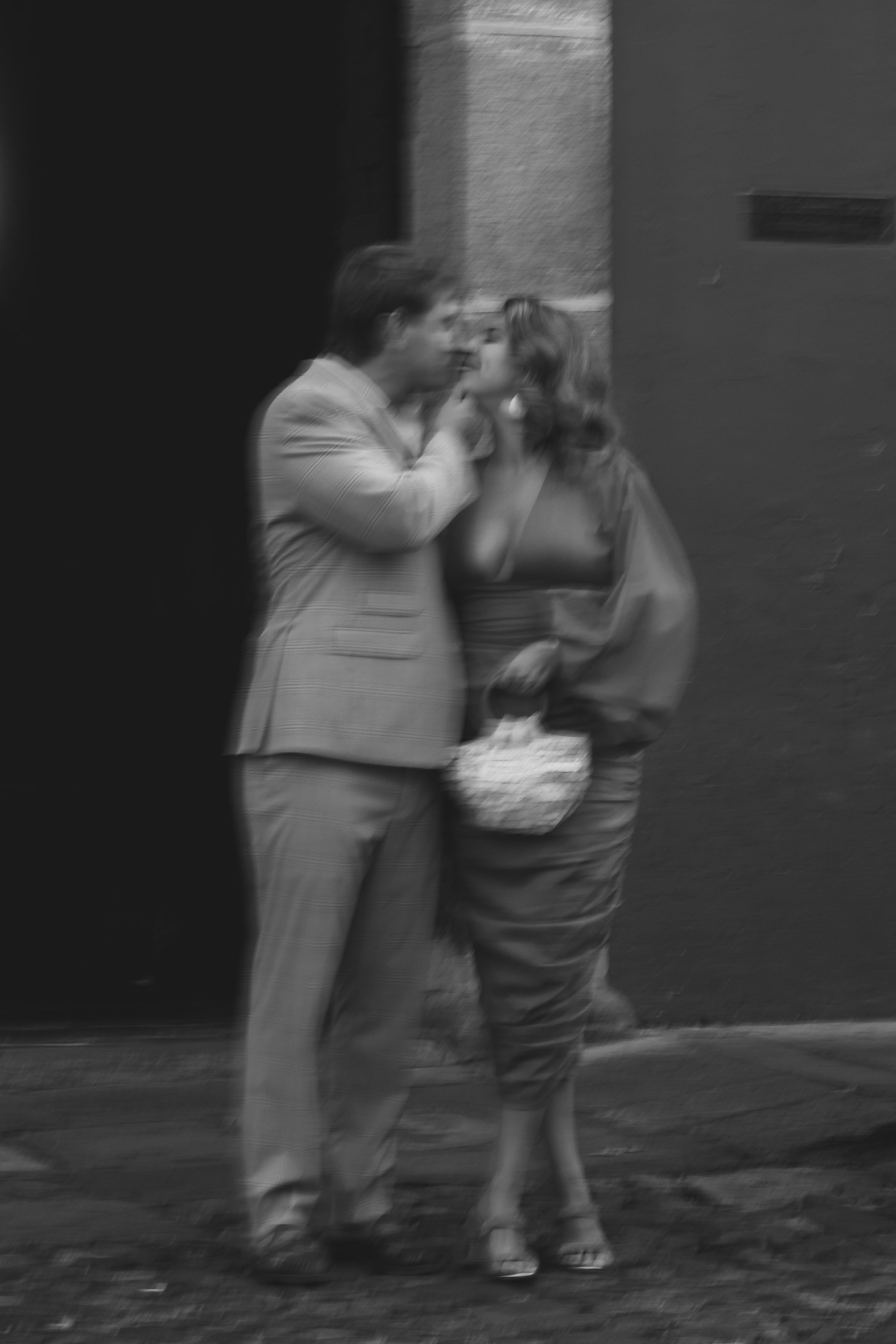 Black and white photo of a man and woman standing close together on a sidewalk, with the man touching the woman's face. The scene appears blurry.