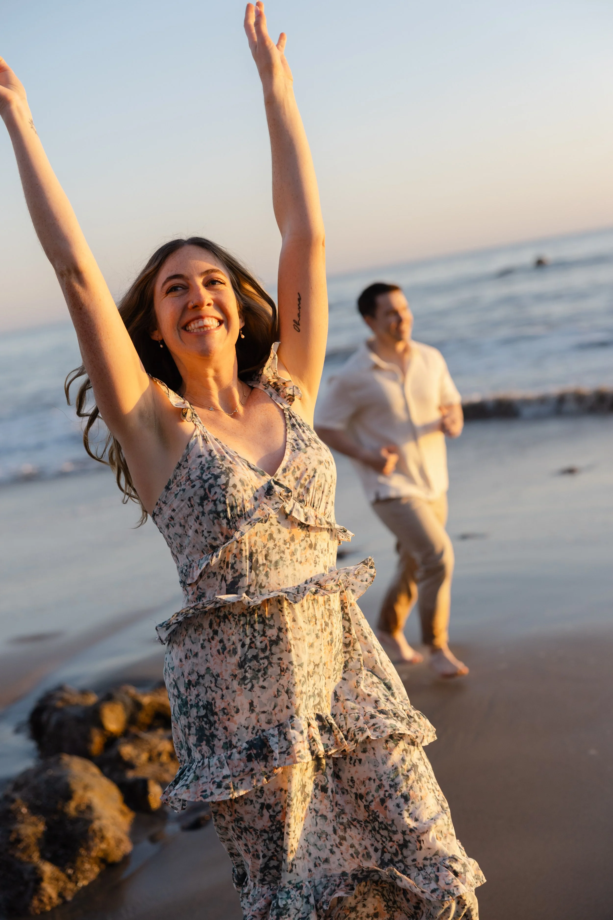 A woman in a floral dress joyfully raises her arms on a beach at sunset, with a man in light-colored clothes dancing in the background.