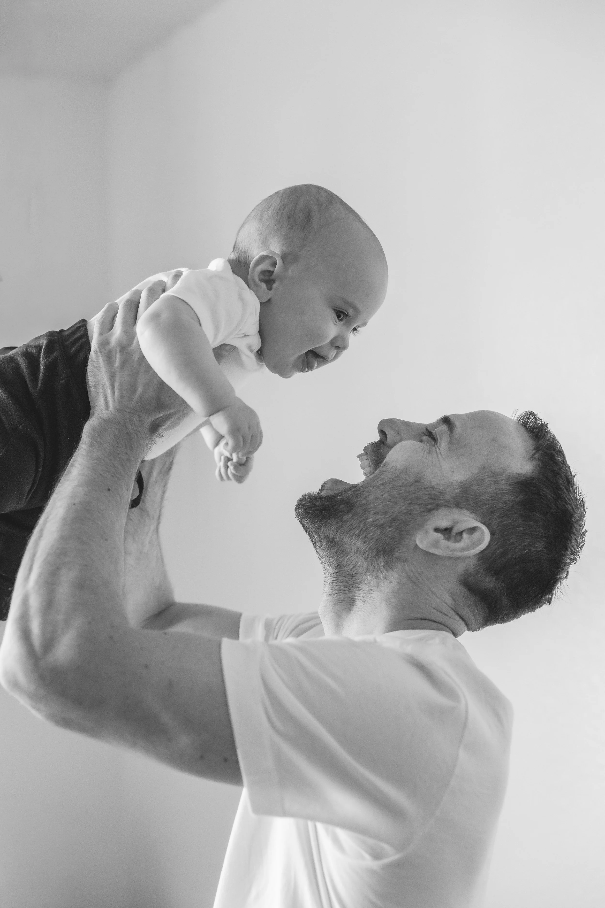 A man lifting a baby boy in the air, both smiling and looking at each other, in a close-up black-and-white photo.