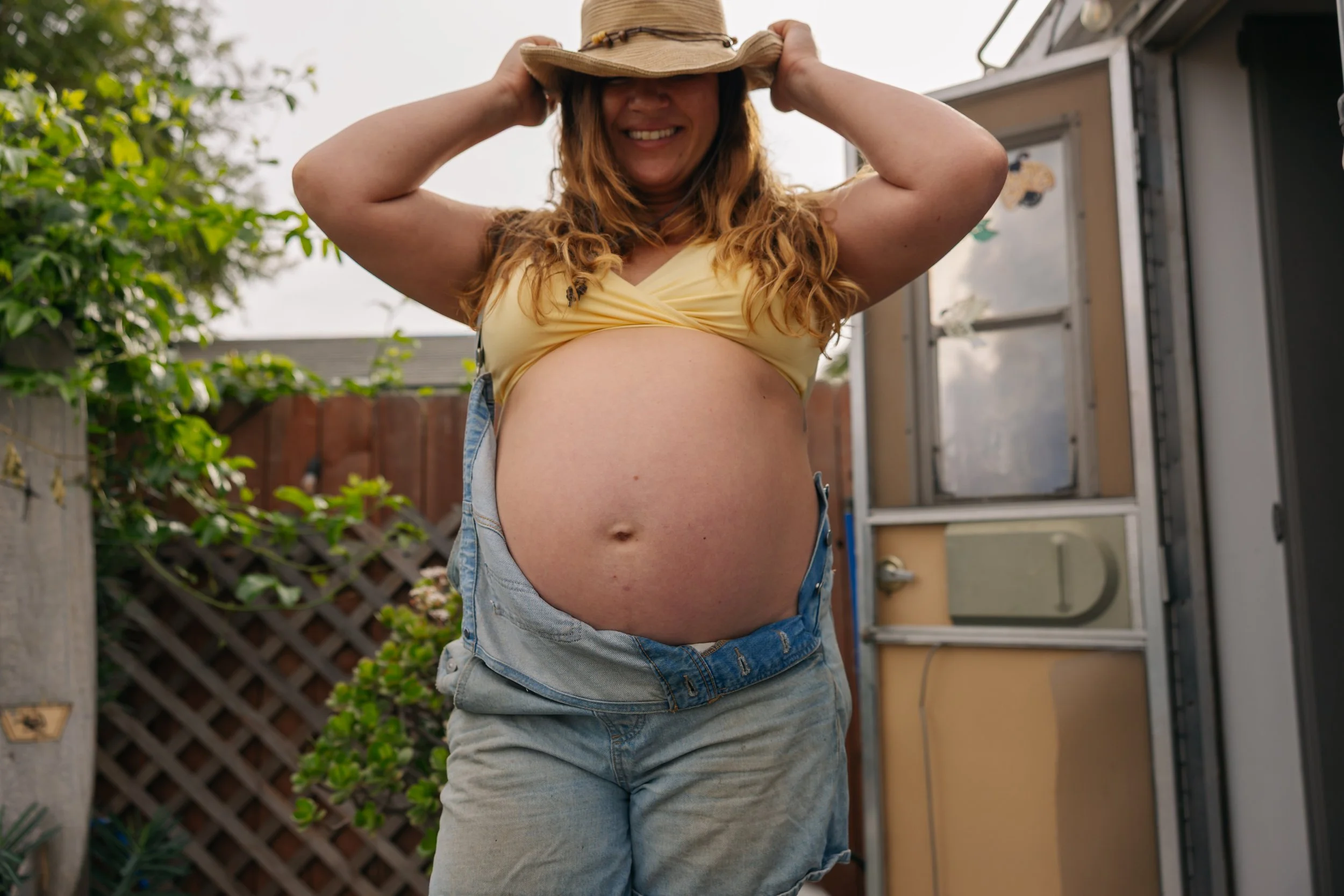 Woman with long red hair smiling, wearing a yellow top and blue jeans, standing outdoors near a shed and greenery, holding a hat above her head.