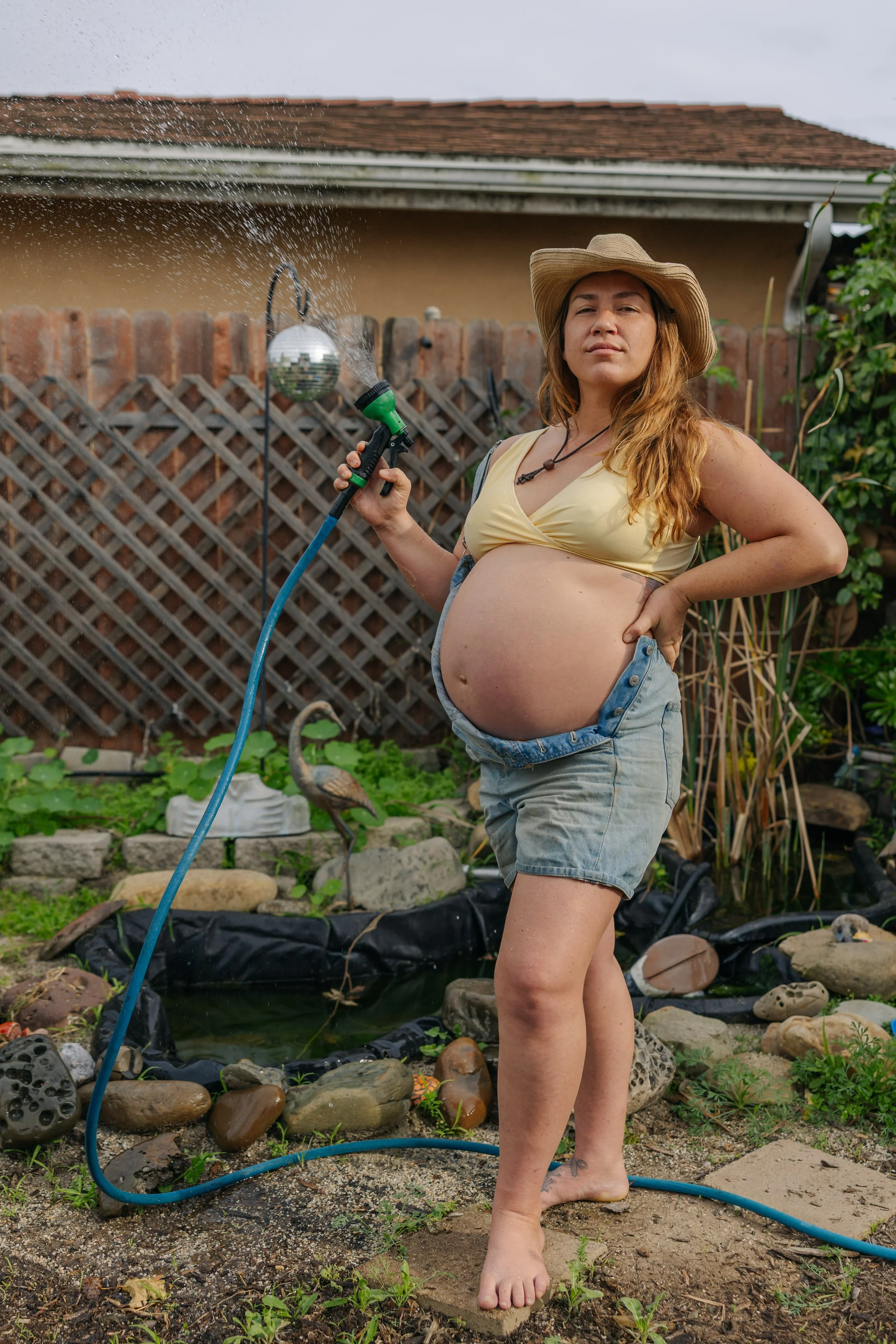 A pregnant woman watering a garden with a hose in a backyard, wearing a straw hat, yellow top, and denim shorts.