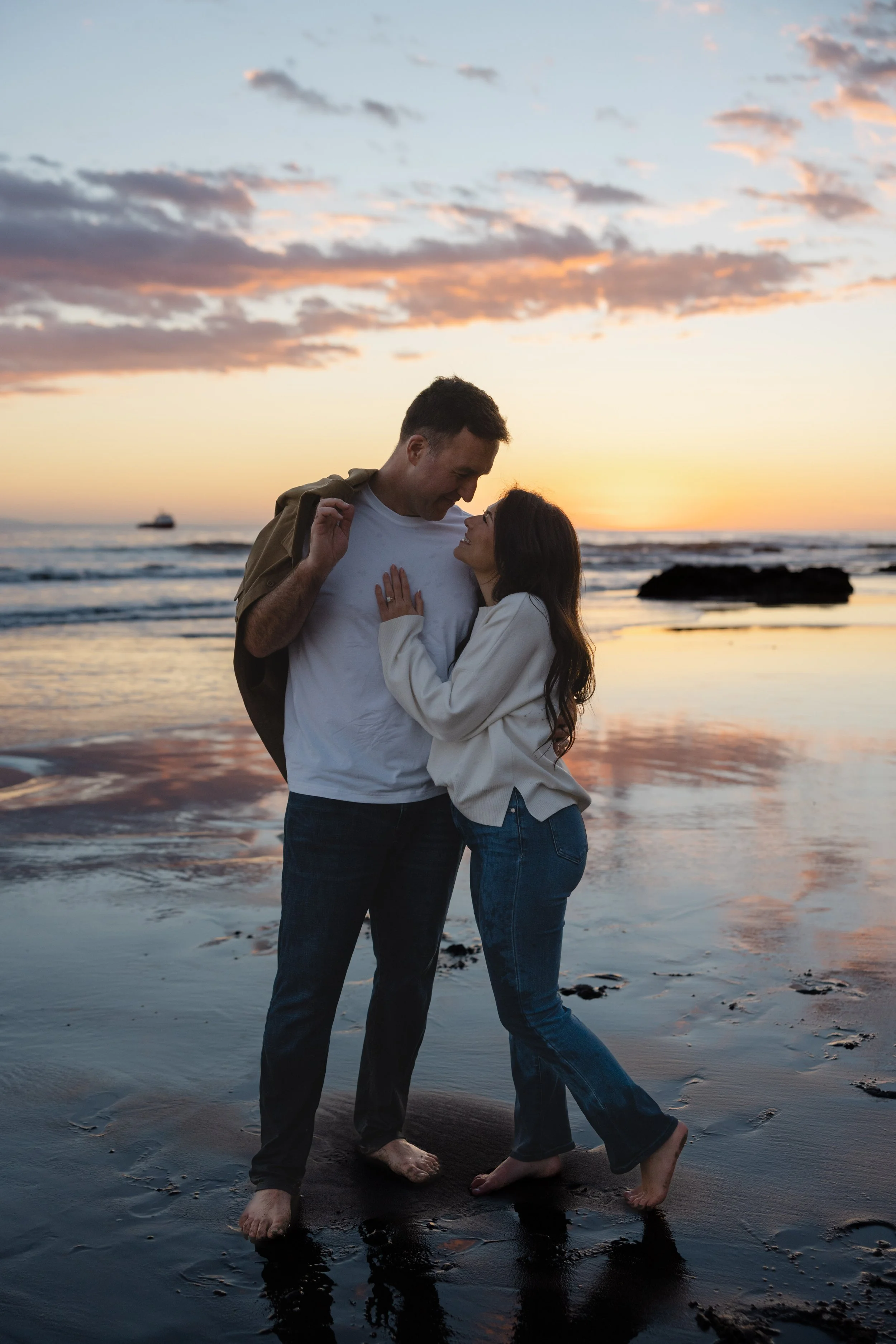 A couple standing close together on a beach at sunset, looking into each other's eyes with the ocean and sky in the background.