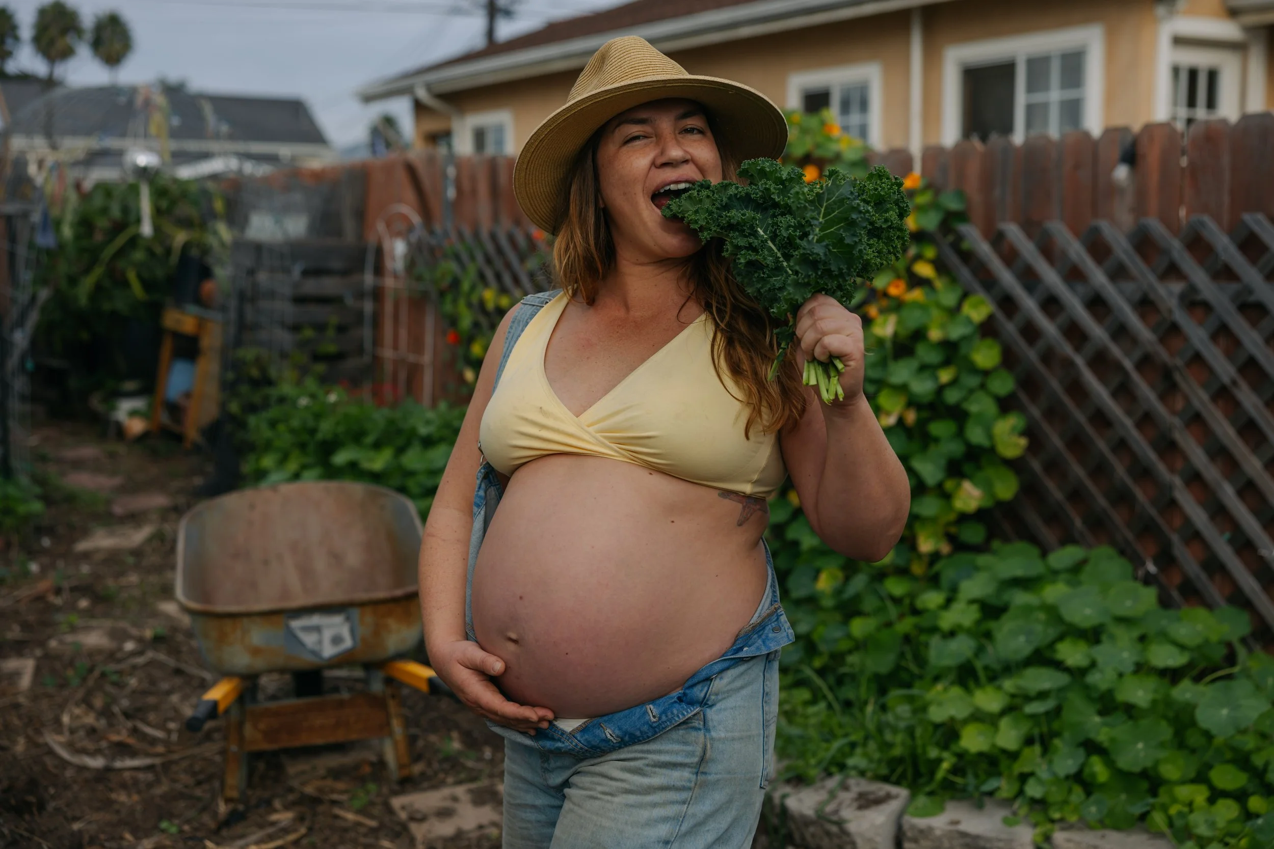 Pregnant woman wearing a straw hat and yellow tank top holding kale in a garden with a wheelbarrow and green plants in the background.