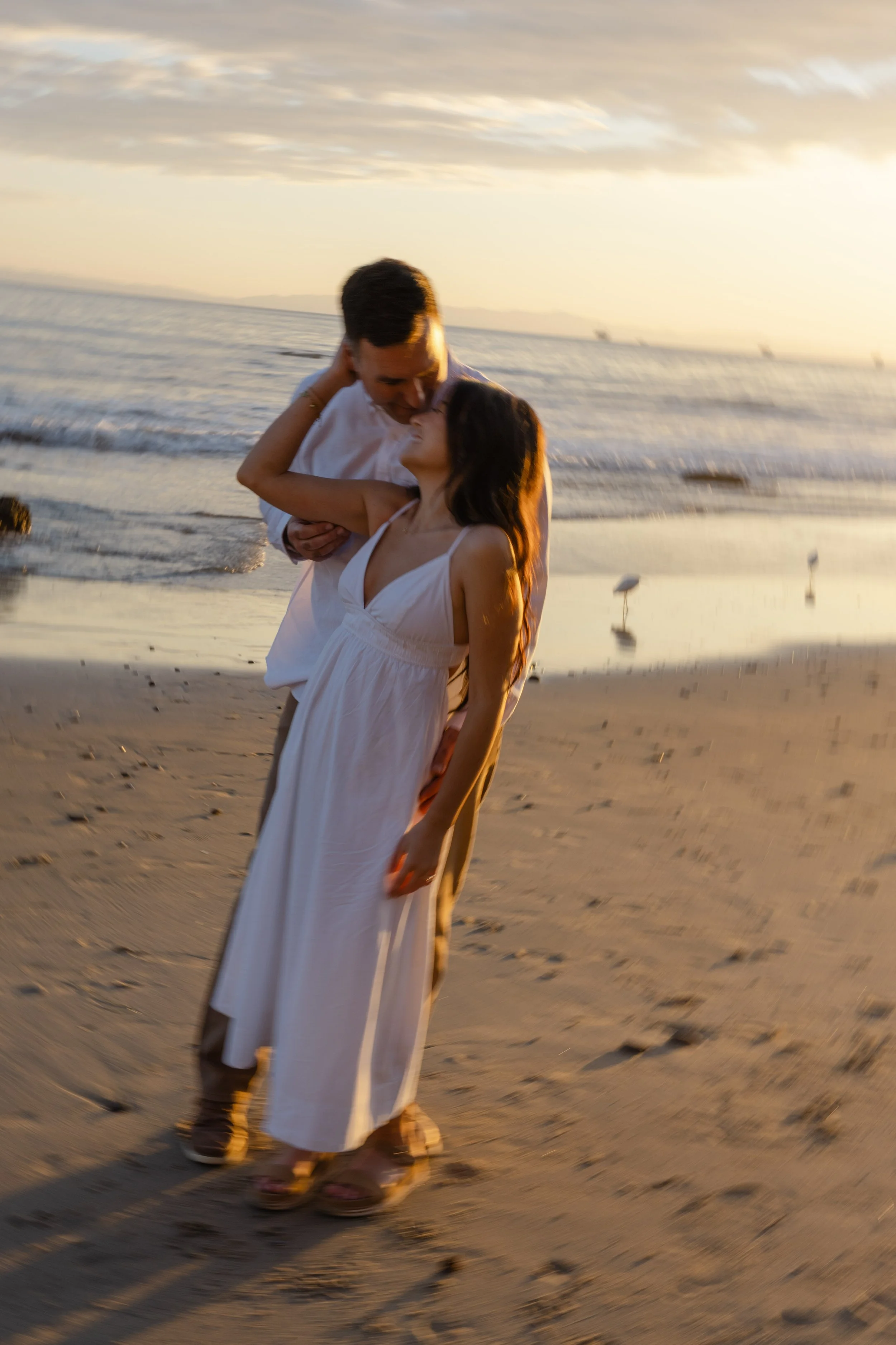 A couple embracing on a beach at sunset, with seagulls in the background and gentle waves