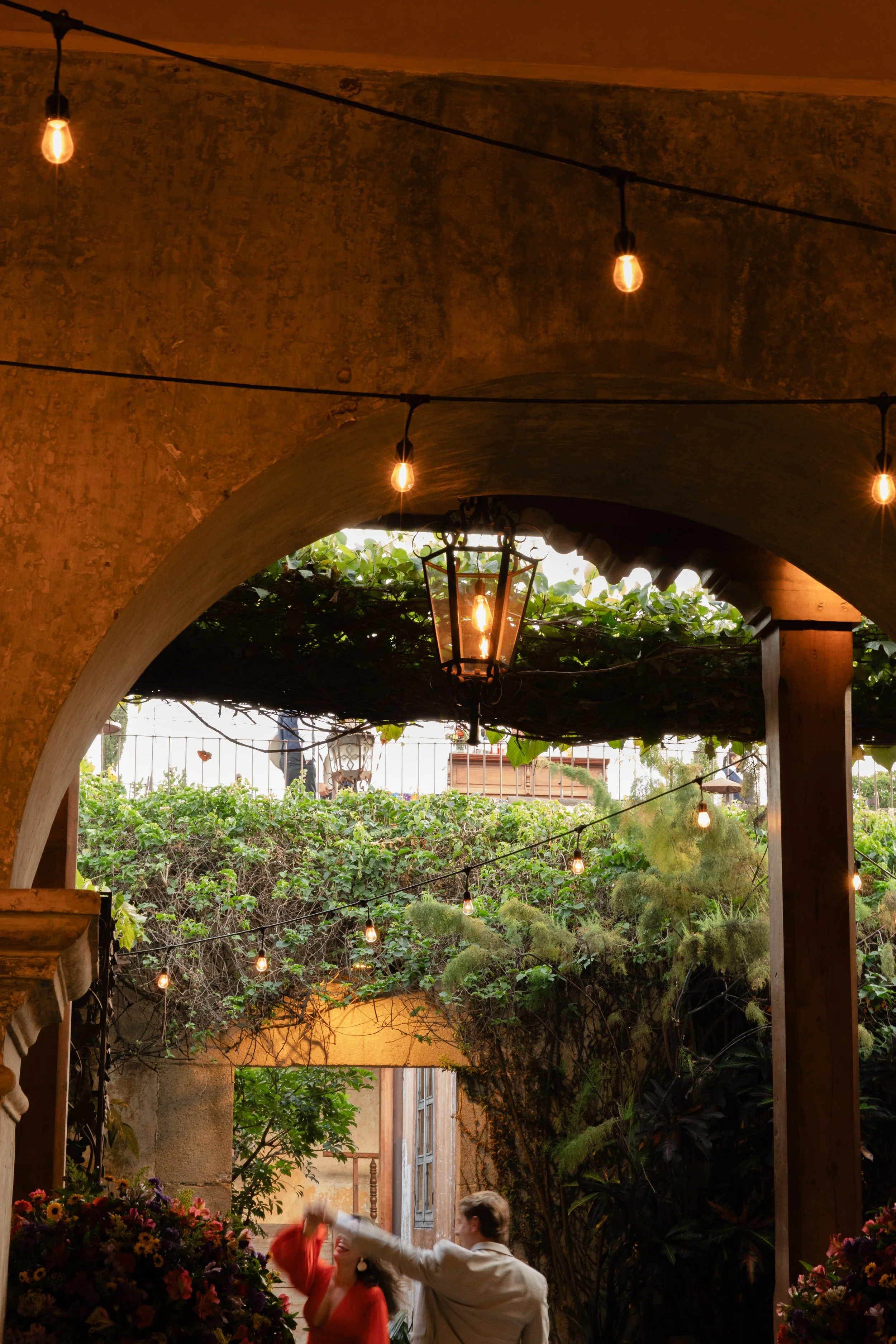 Two people dancing or playing under a pergola decorated with lush greenery, string lights, and a hanging lantern in an outdoor patio area.