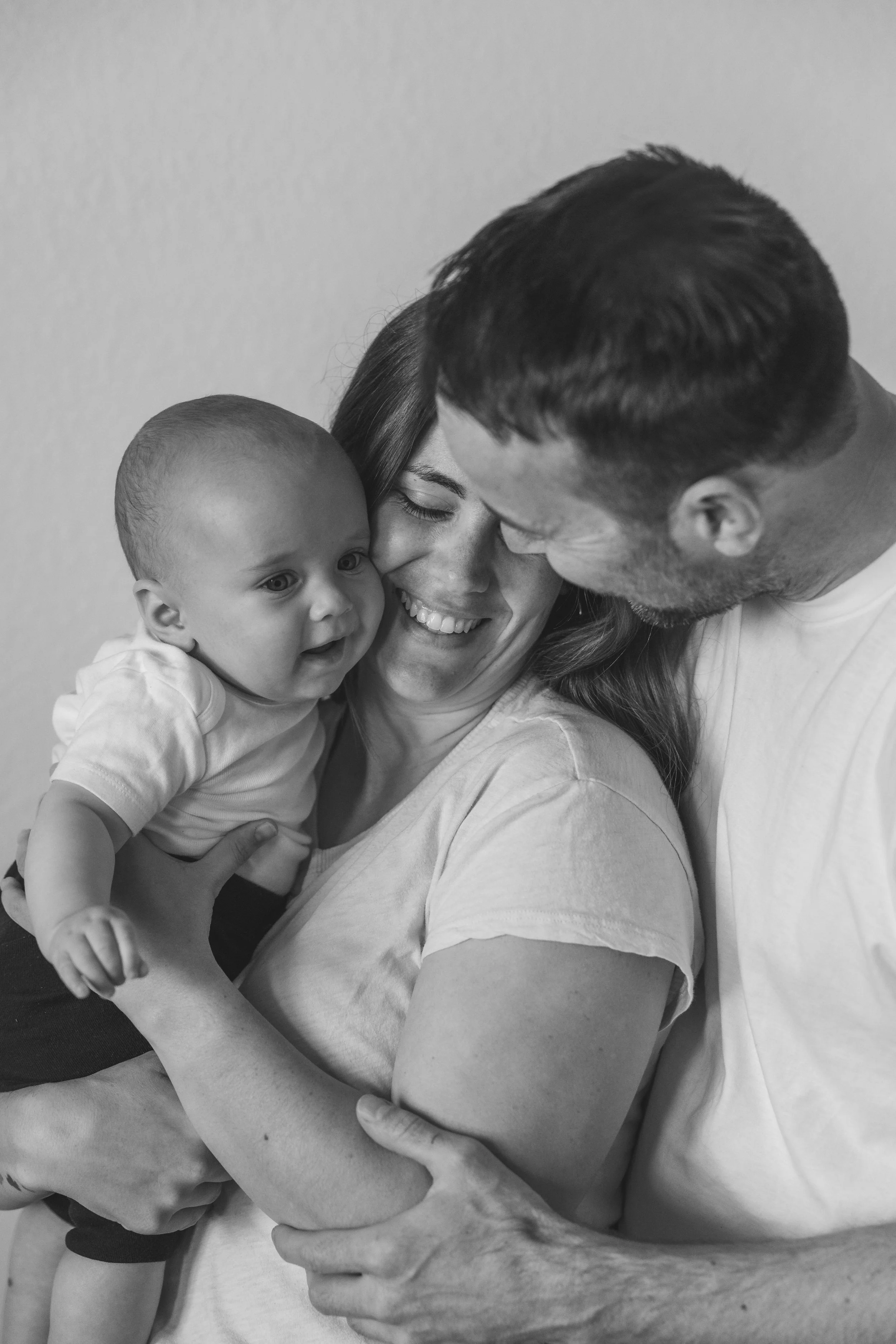 A black and white photo of a happy family, including a woman holding a baby, with a man leaning close to them, all smiling and loving.