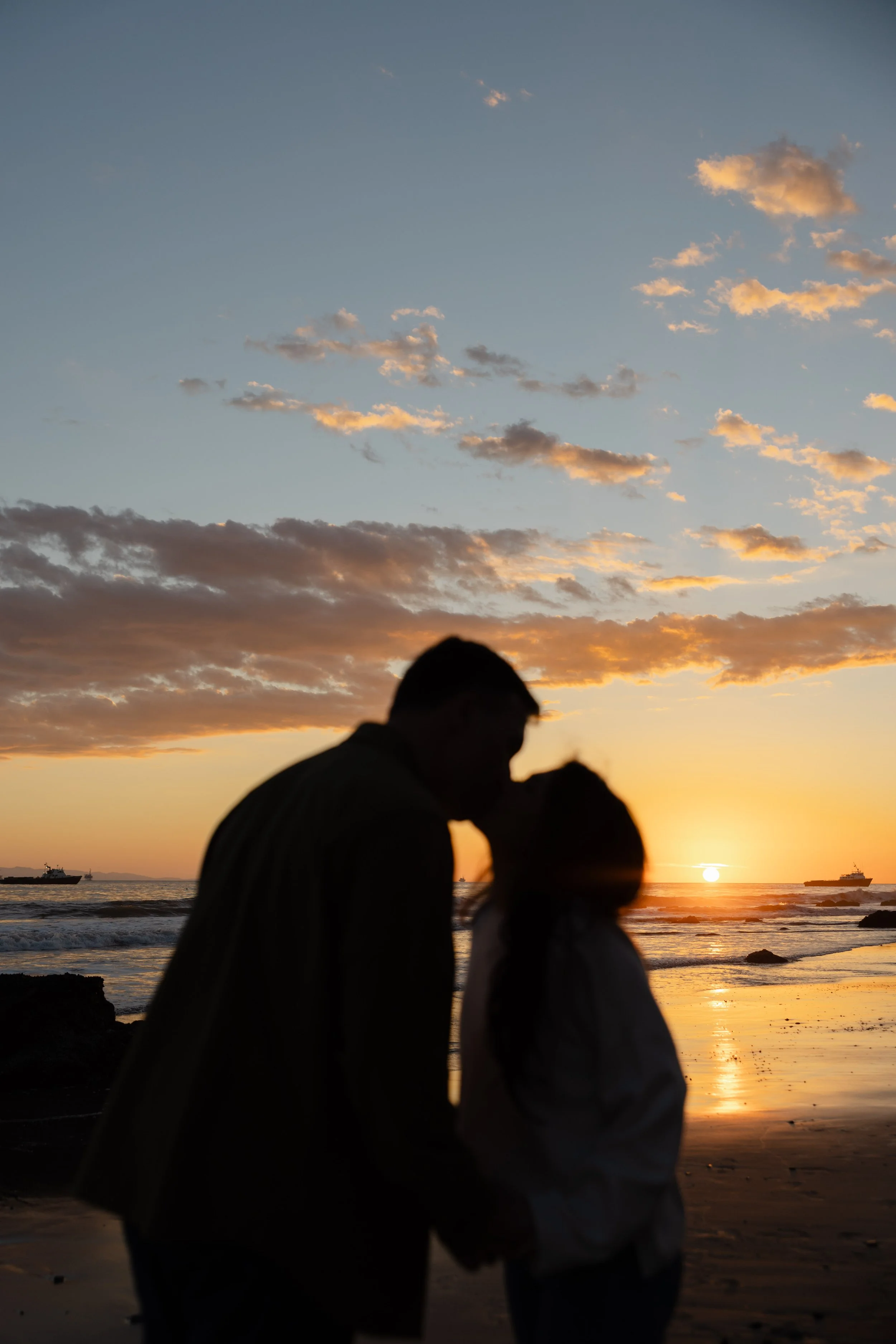 Silhouette of a couple kissing on the beach at sunset with the ocean and boats in the background.