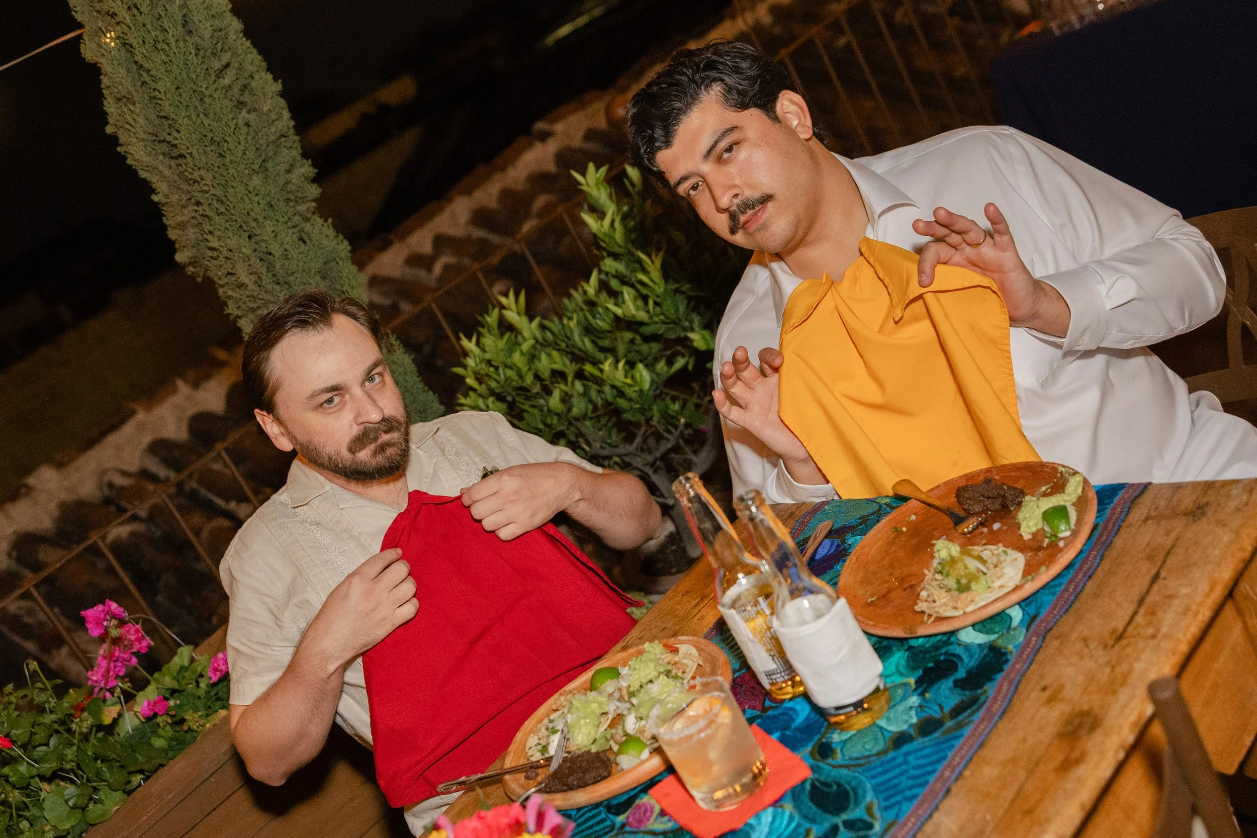 Two men sitting at a wooden table with food, drinks, and napkins in an outdoor setting at night. One man is wearing a beige shirt and a red apron, holding his apron, and the other man is wearing a white shirt with an orange apron, with food on a plat