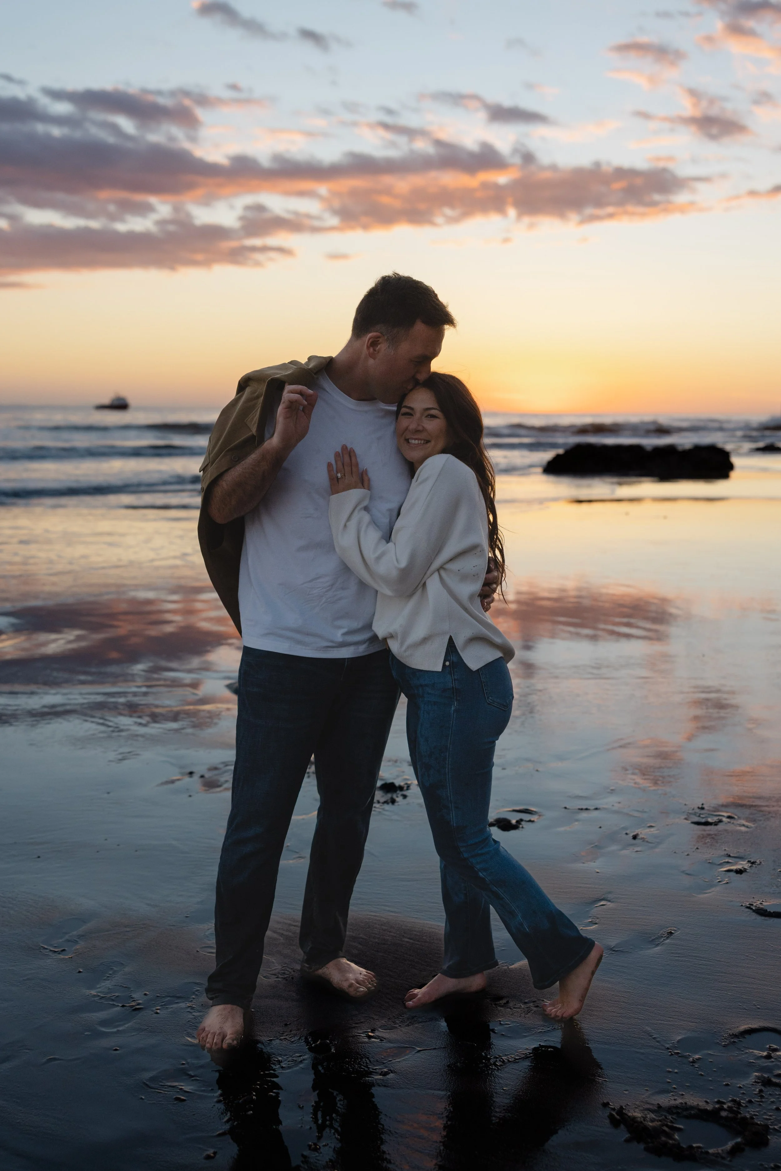 A couple standing barefoot on the beach during sunset, embracing and smiling, with the ocean and rocks in the background.