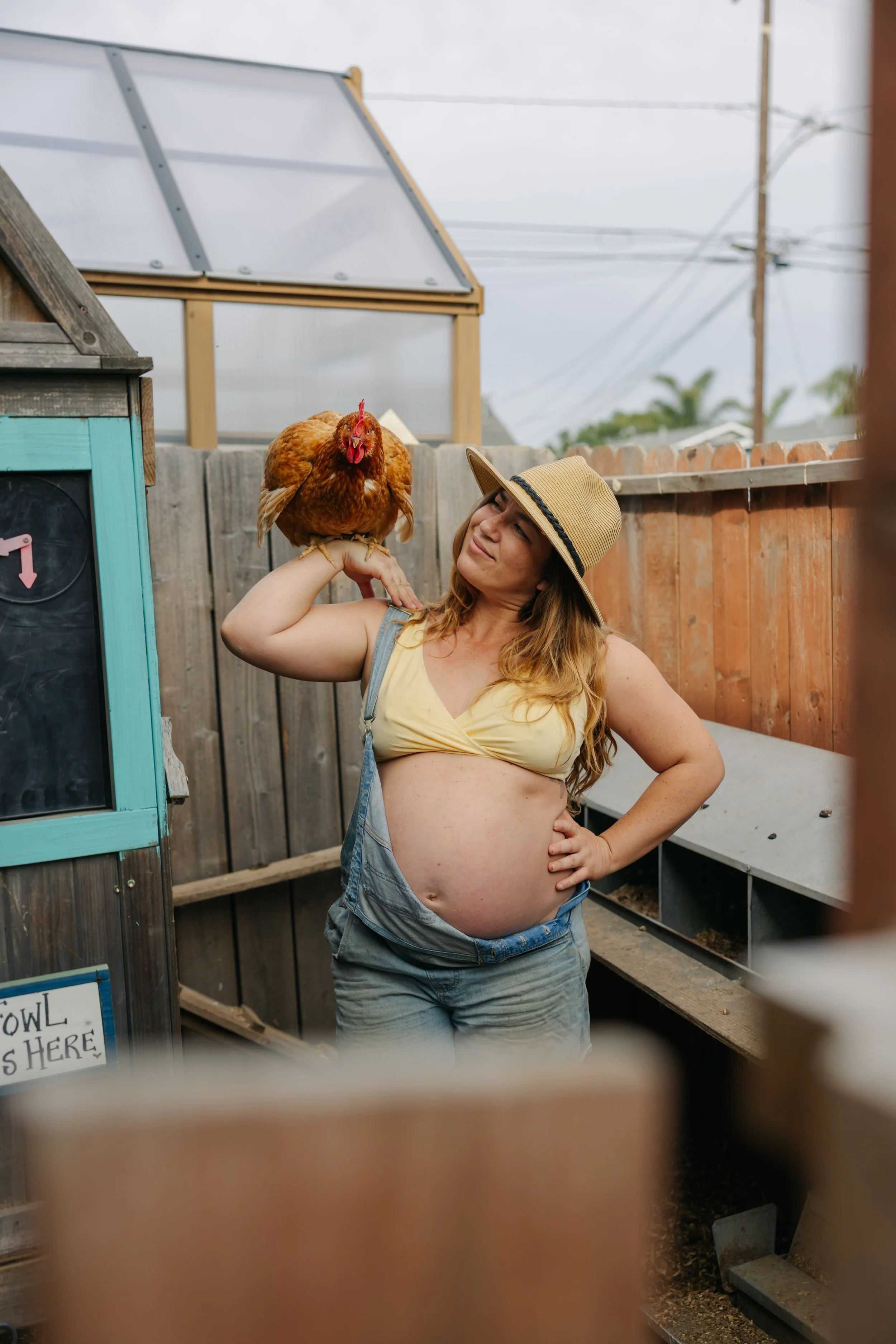 A pregnant woman with long red hair, wearing a yellow top, denim overalls, and a straw hat, is holding a hen on her shoulder, standing in a fenced outdoor area with a greenhouse and a sign that says 'BOWL'. She is smiling and looking at the hen.