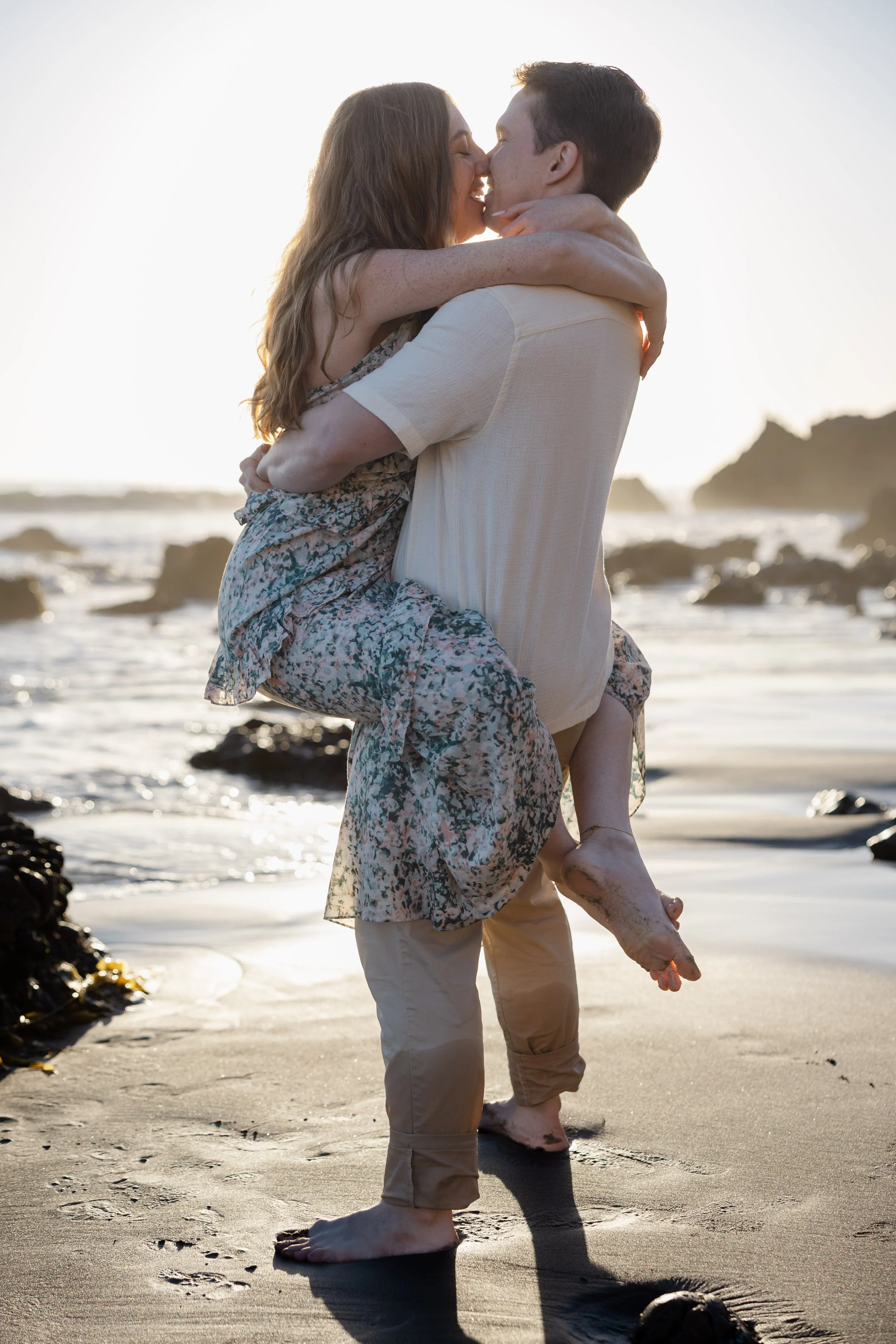A couple is embracing on the beach at sunset, with the woman in a floral dress being held by the man as they share a kiss.