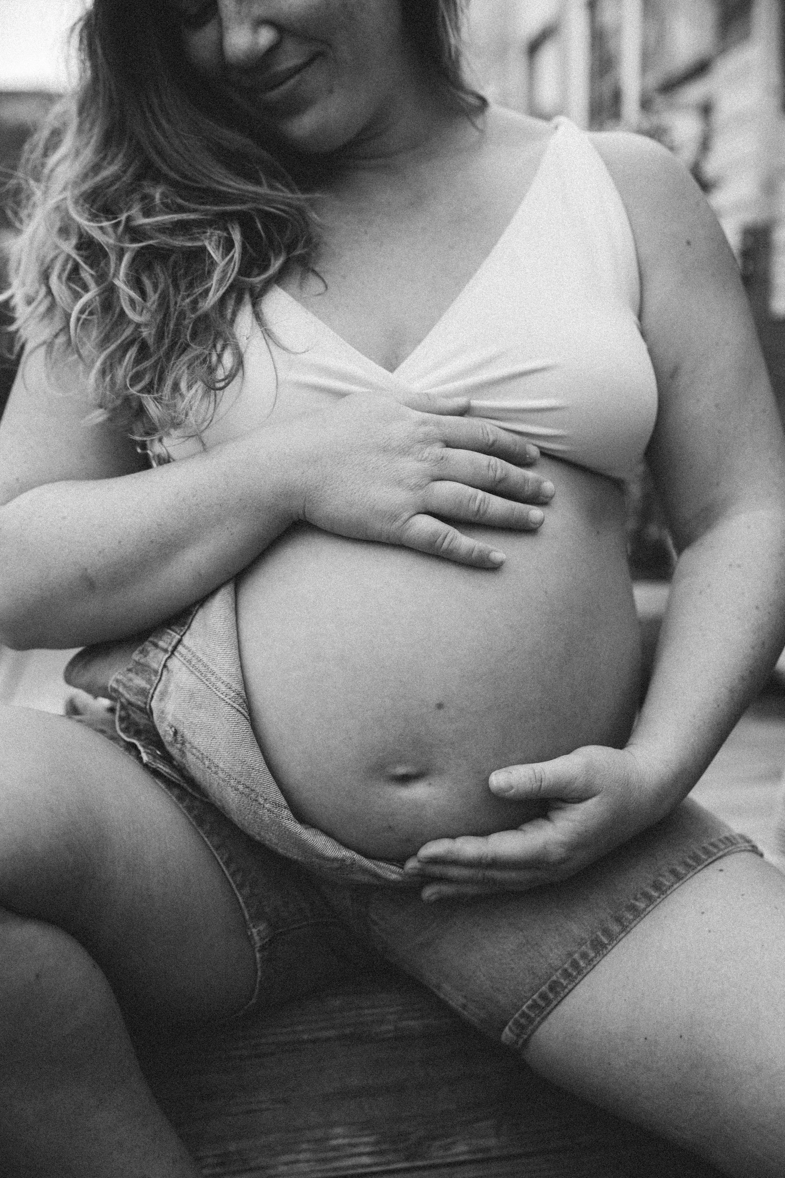 A pregnant woman is sitting with her legs apart, cradling her belly with her hands. She is smiling and wearing a sleeveless top and denim shorts.