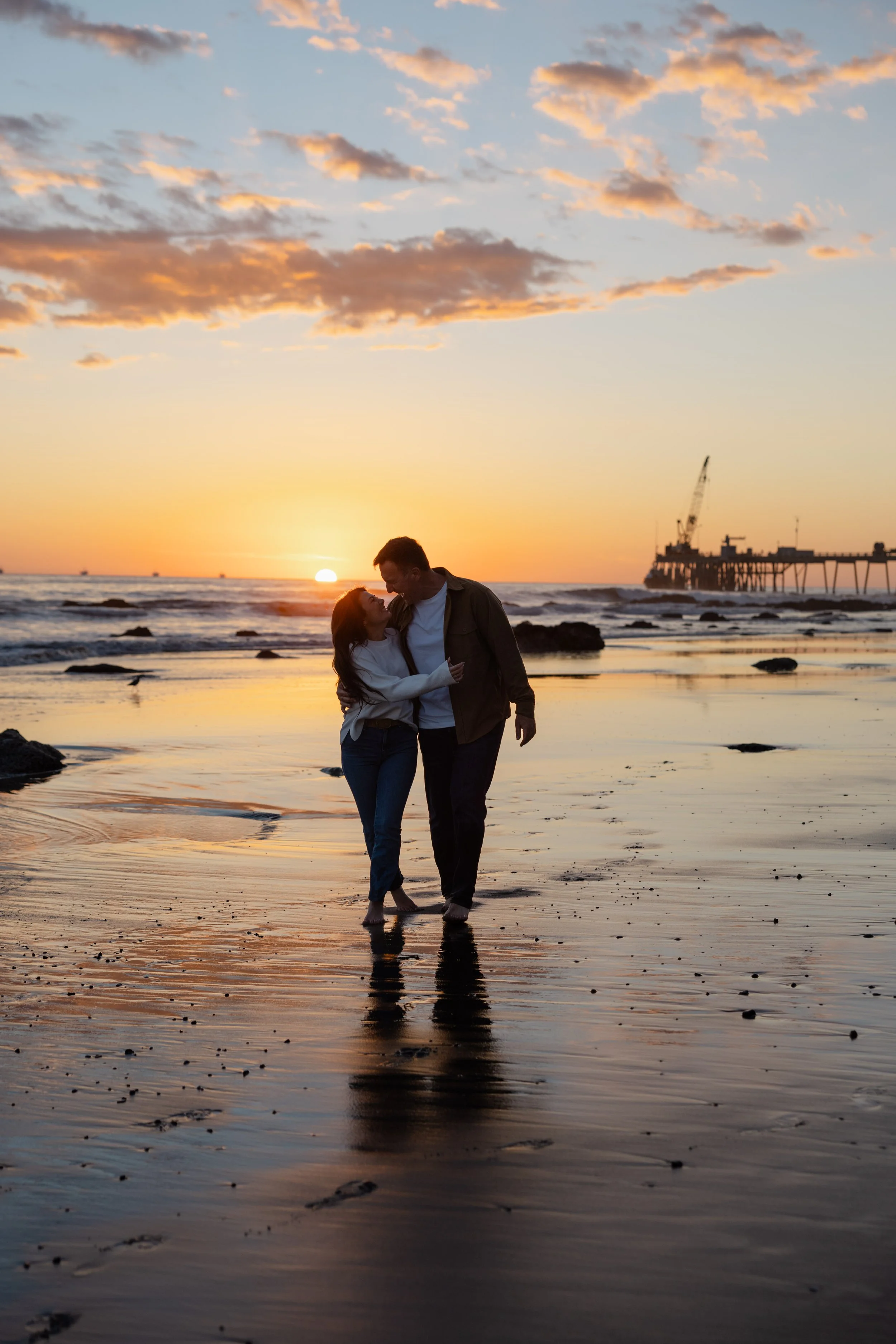 A couple walking barefoot on a beach at sunset, with the ocean and a pier with construction equipment in the background.