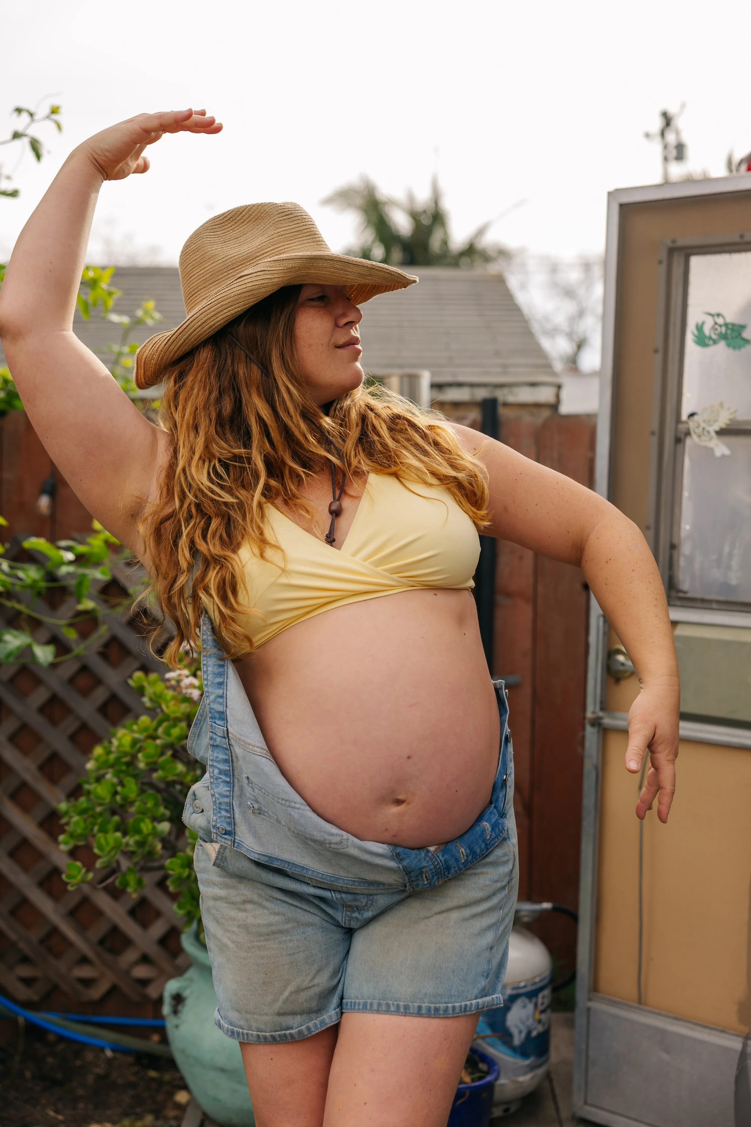 A woman with red hair wearing a large sun hat, a yellow crop top, and denim shorts, striking a dance pose outdoors.