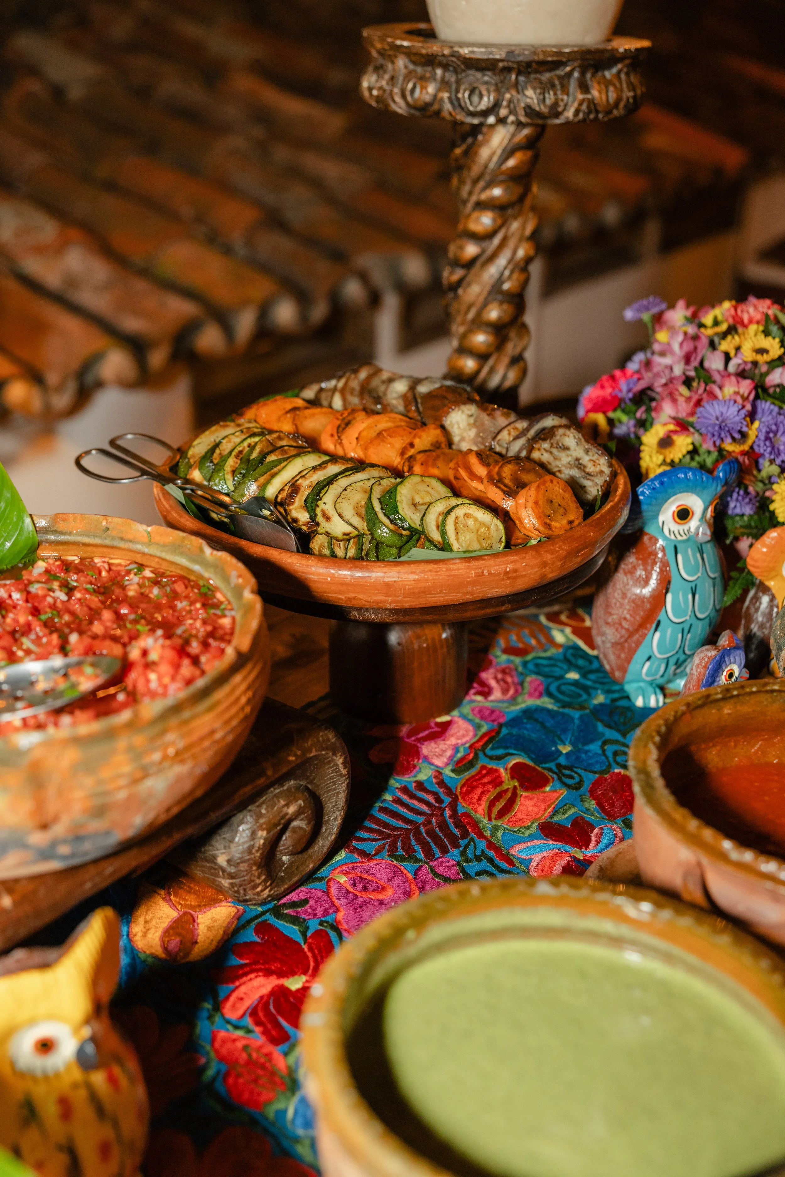 A colorful table setting featuring a platter of grilled vegetables including zucchini, carrots, and onions, and a bowl of salsa, with vibrant Mexican-style pottery and a floral centerpiece.