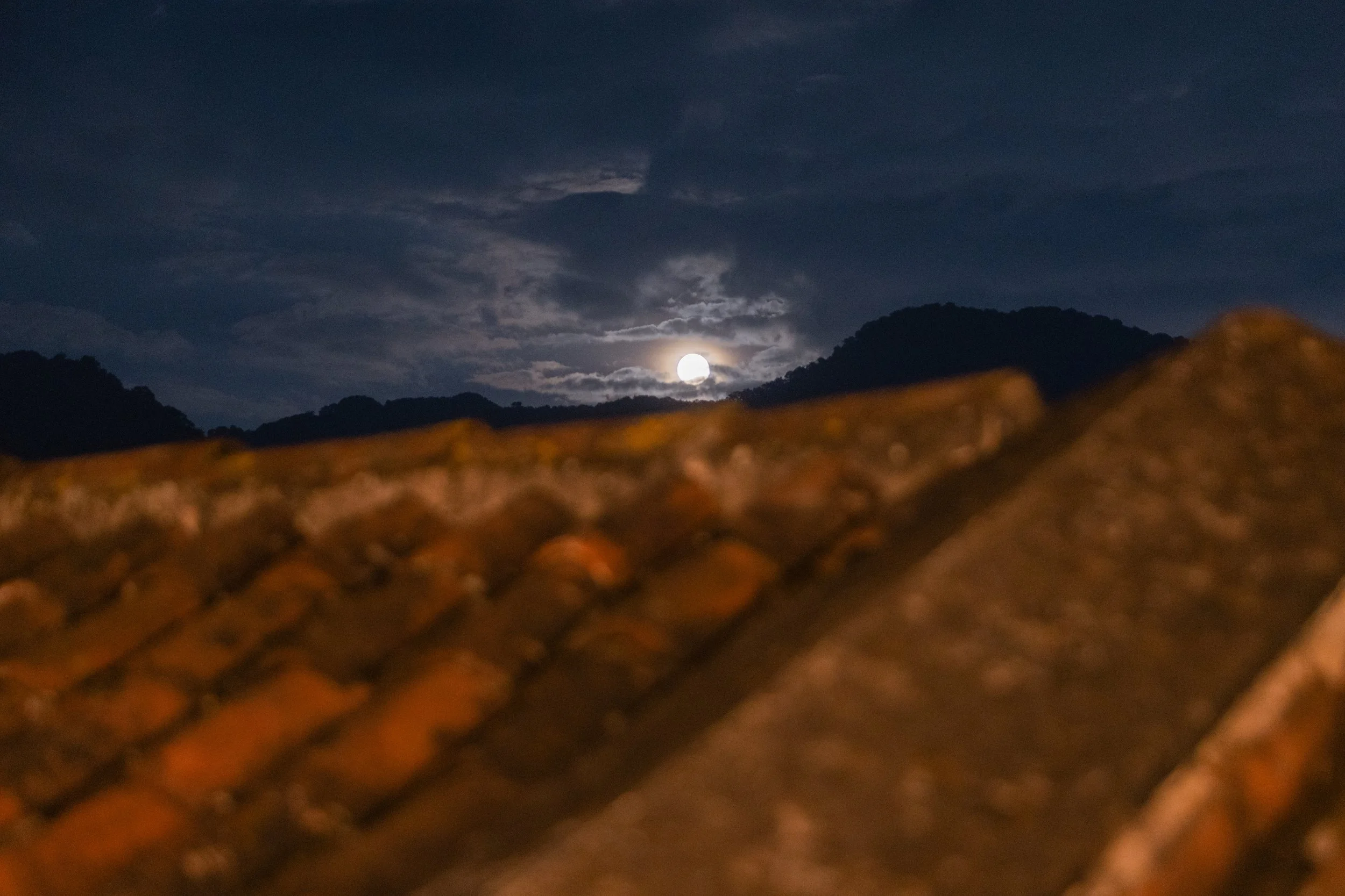 Nighttime scene with a bright full moon rising above mountains and clouds, partially obscured by rooftop tiles in the foreground.