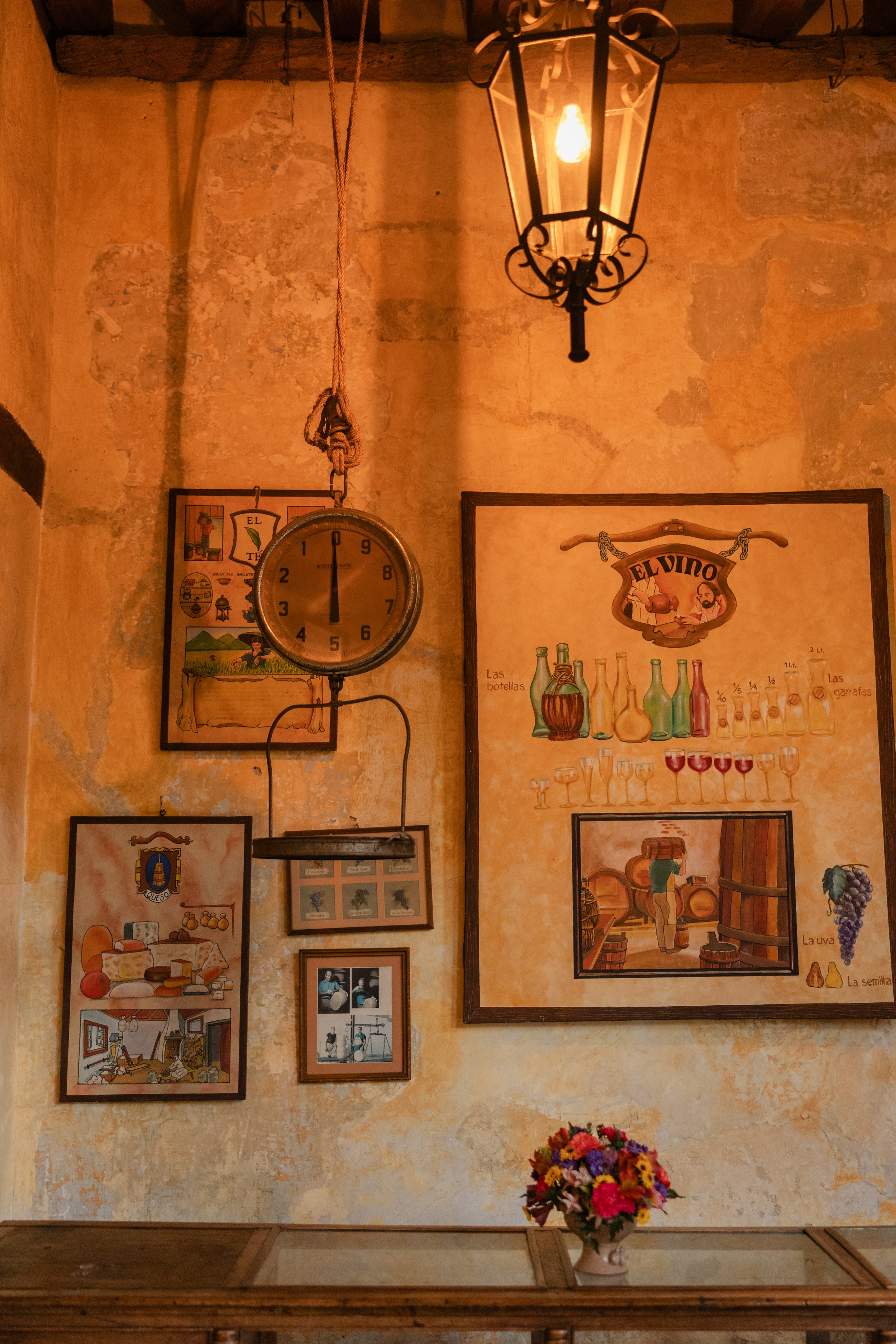 Interior of a rustic restaurant with a vintage clock hanging on the wall, surrounded by colorful framed pictures, with a hanging lantern light above and a vase of flowers on a wooden table in the foreground.