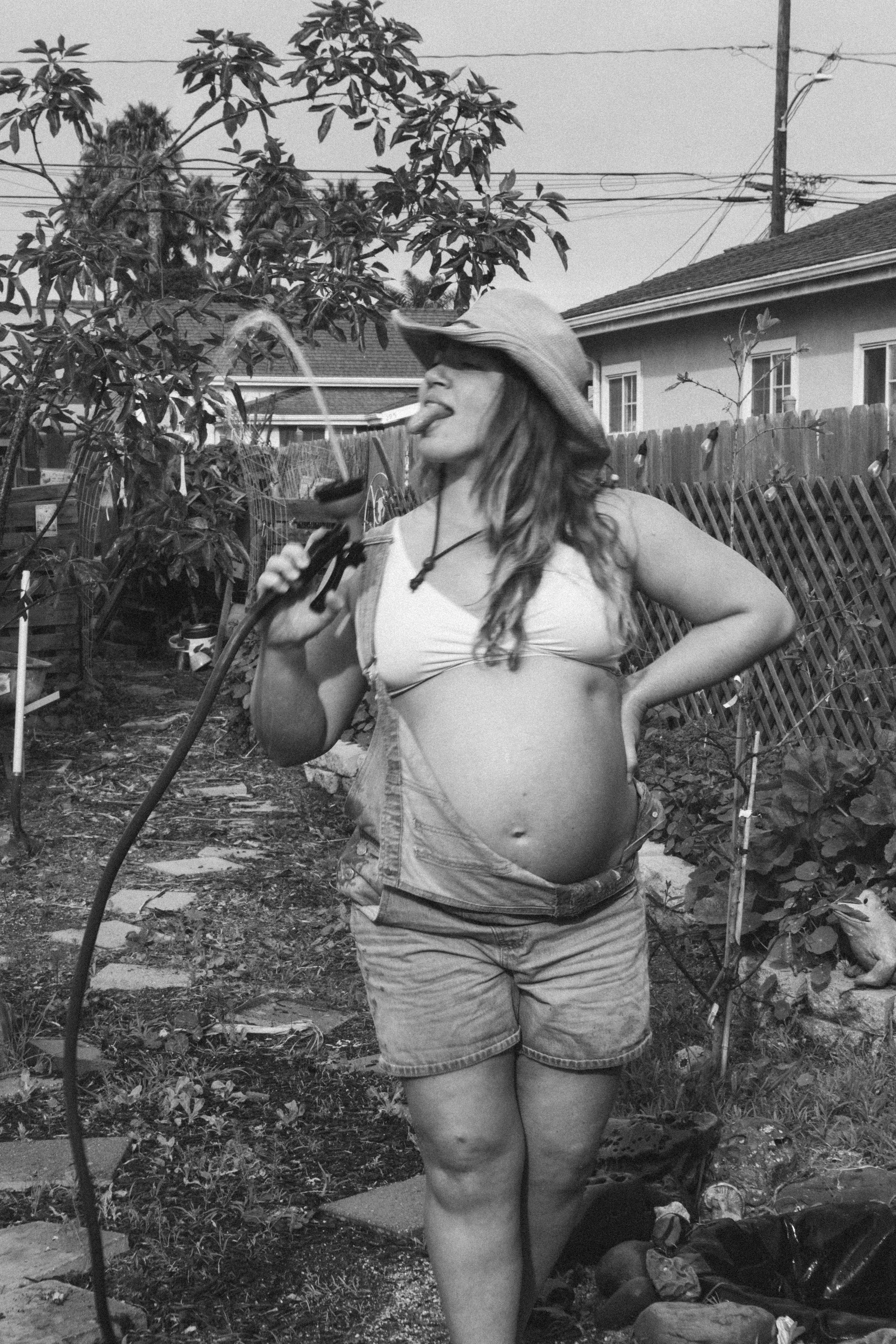 A pregnant woman wearing a sun hat and shorts in a garden, holding a garden hose and playing with water, sticking her tongue out.