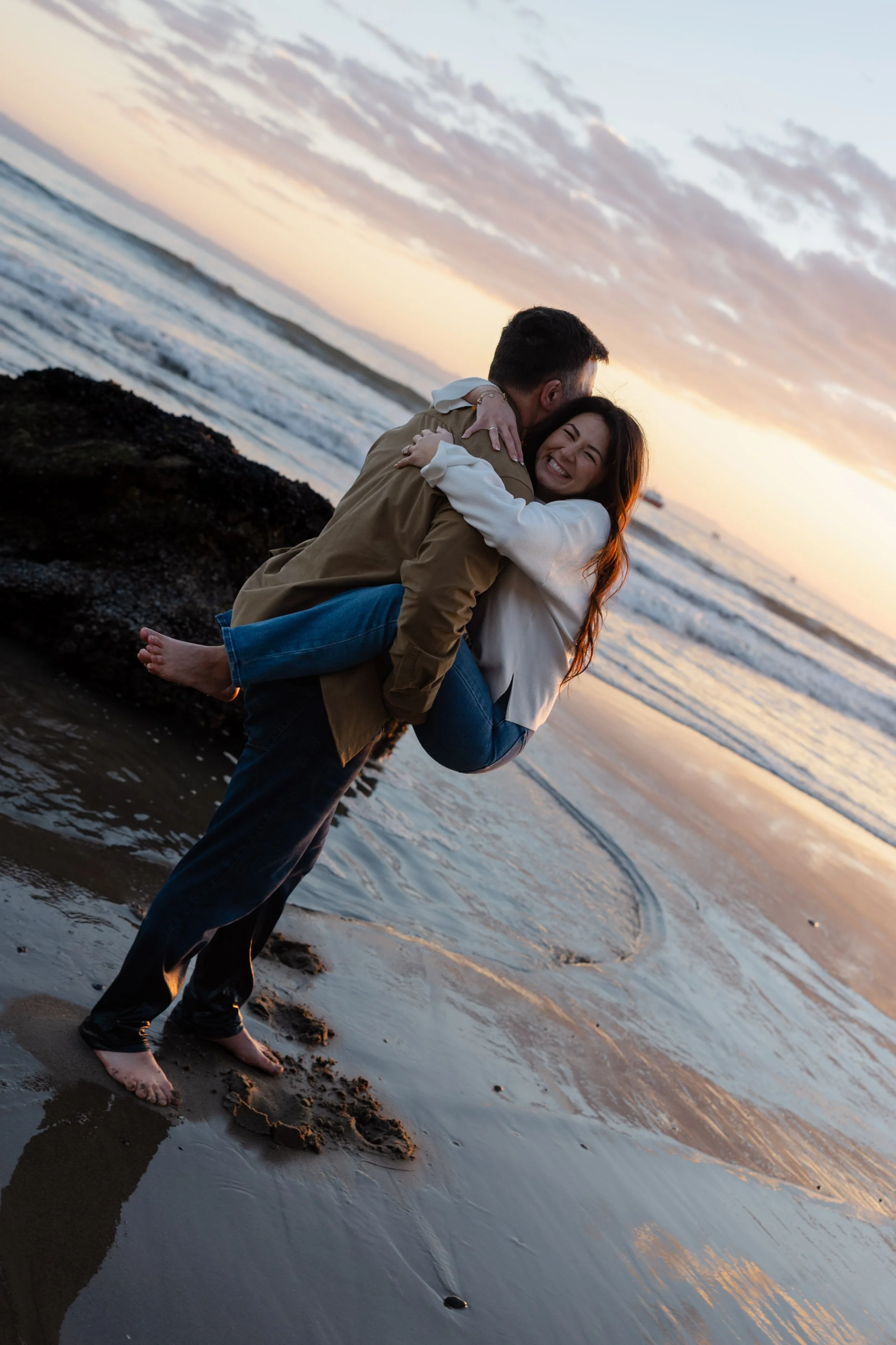 A couple hugging on a beach at sunset, with the man lifting the woman in his arms, both smiling happily.