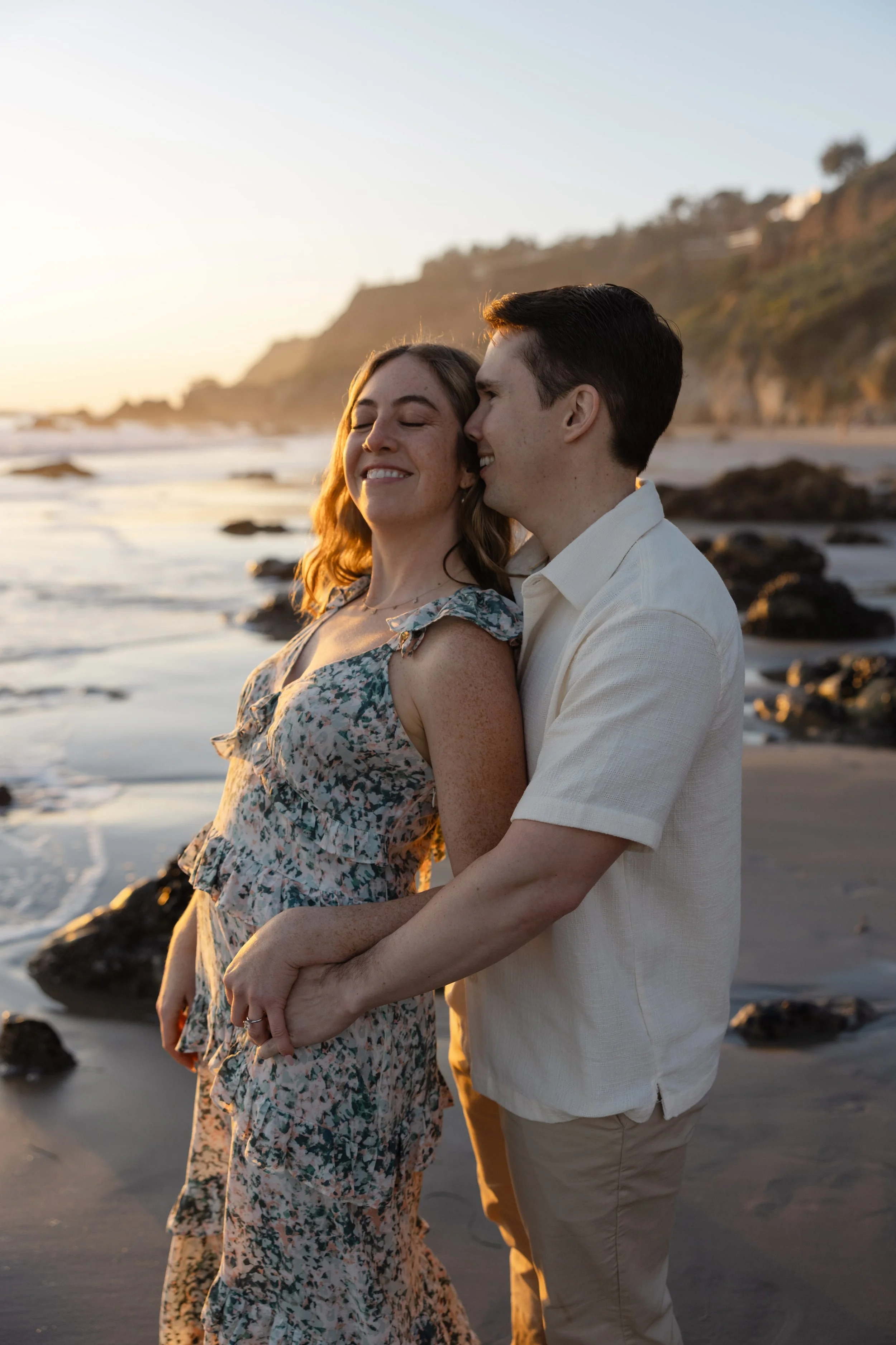 A couple holding hands and smiling at each other on the beach at sunset, with rocky shoreline and hills in the background.