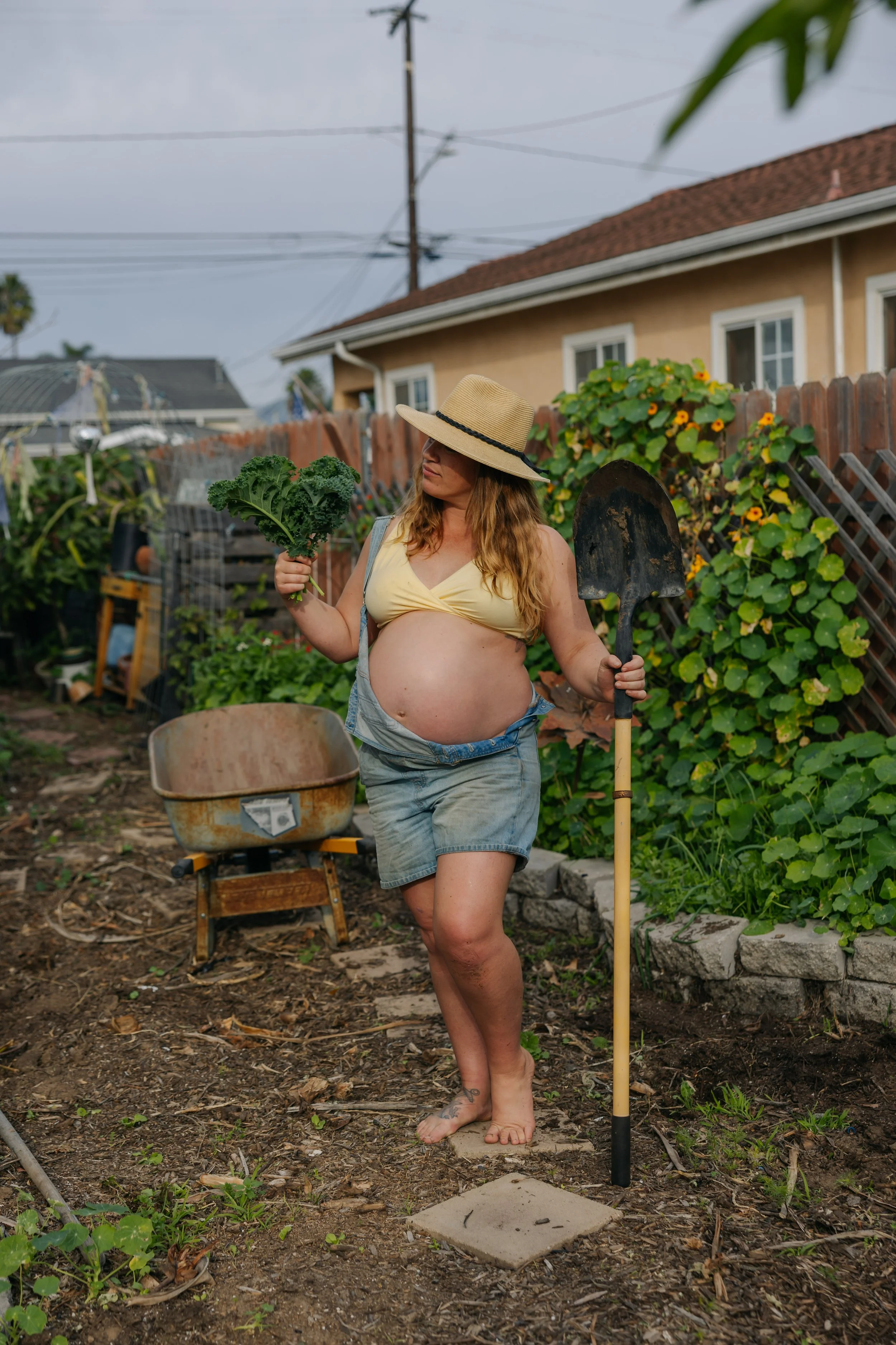 A pregnant woman outdoors in her garden, holding a leafy green vegetable in one hand and a gardening shovel in the other, wearing a straw hat, a yellow top, and denim shorts.