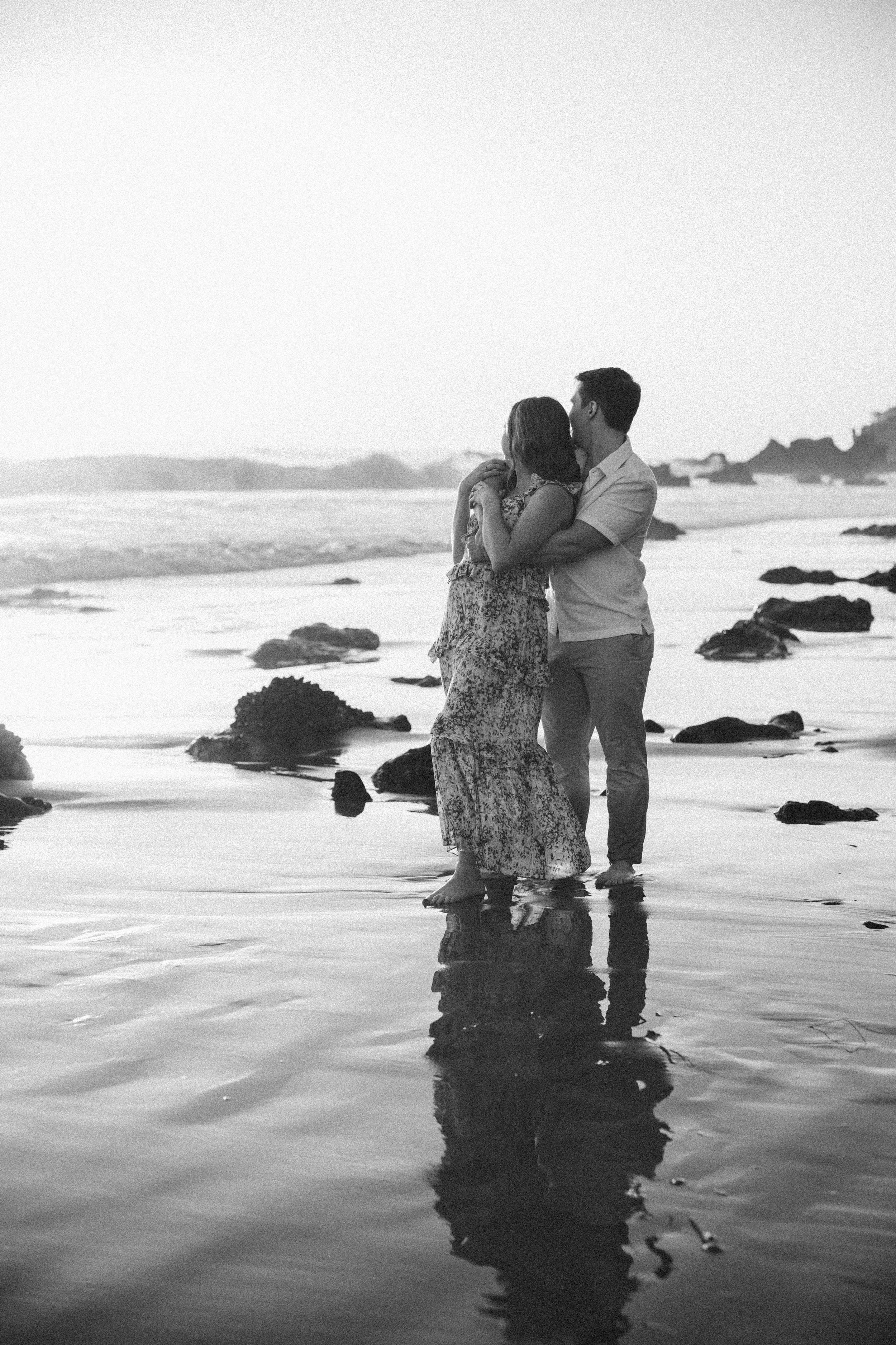 A black and white photo of a couple embracing on the beach, with rocks and ocean waves in the background during sunset.