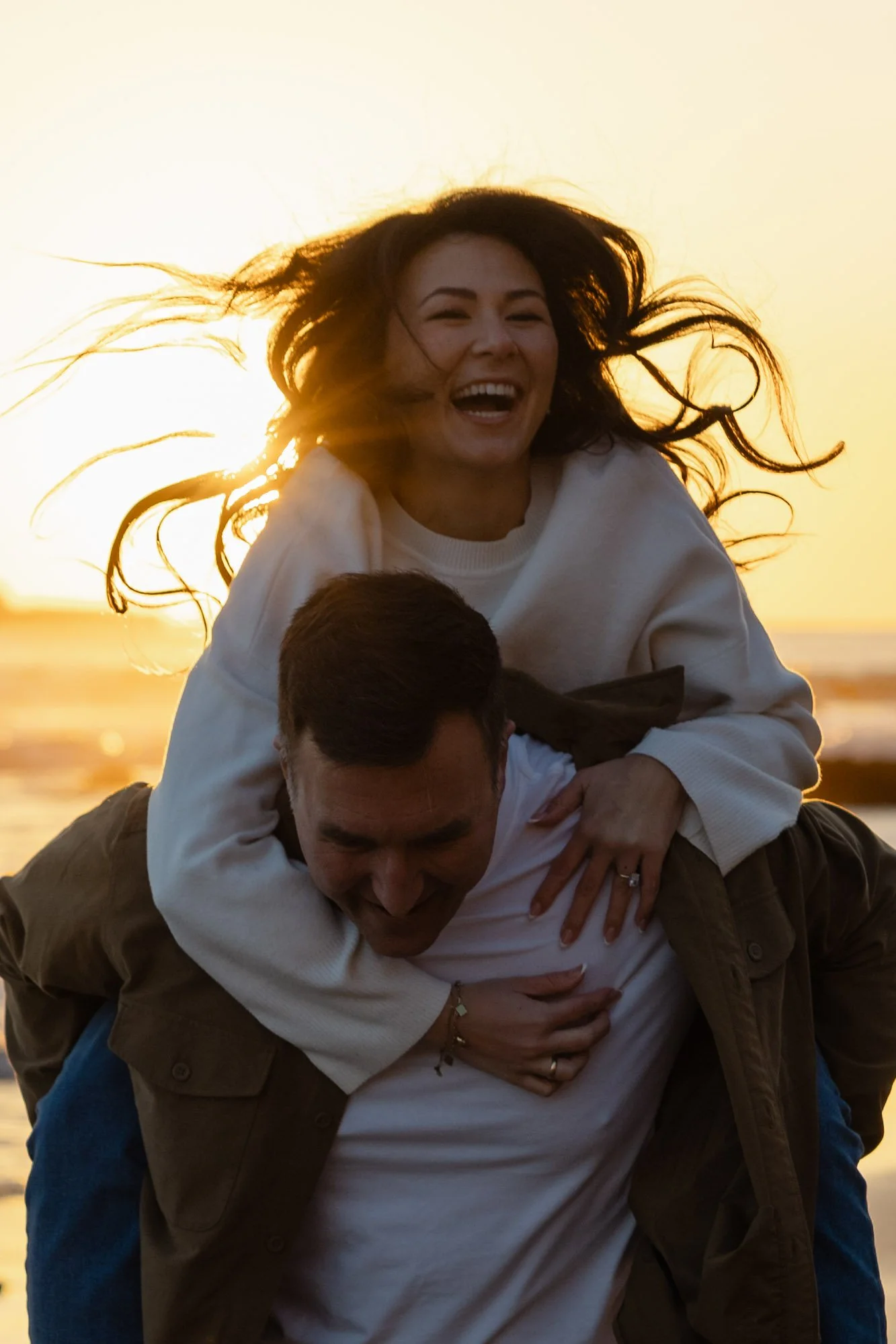 A woman laughing and smiling while sitting on a man's shoulders at the beach during sunset, with the sun behind her hair.