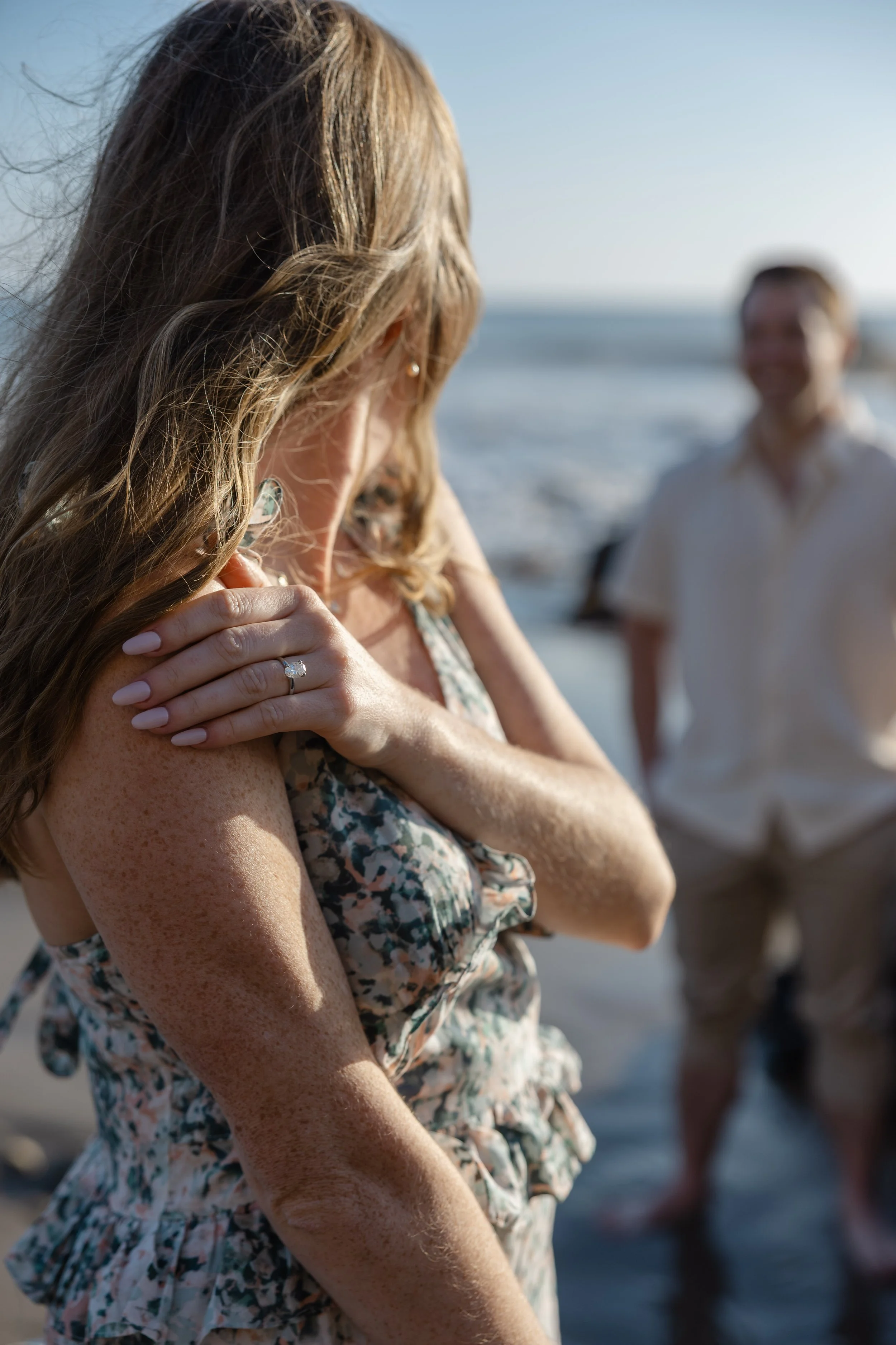 A woman with freckles and wavy hair, wearing a floral dress, stands on the beach with her hand on her shoulder, and is showing an engagement ring on her finger. In the background, a man in a white shirt and another person are near the water, out of f