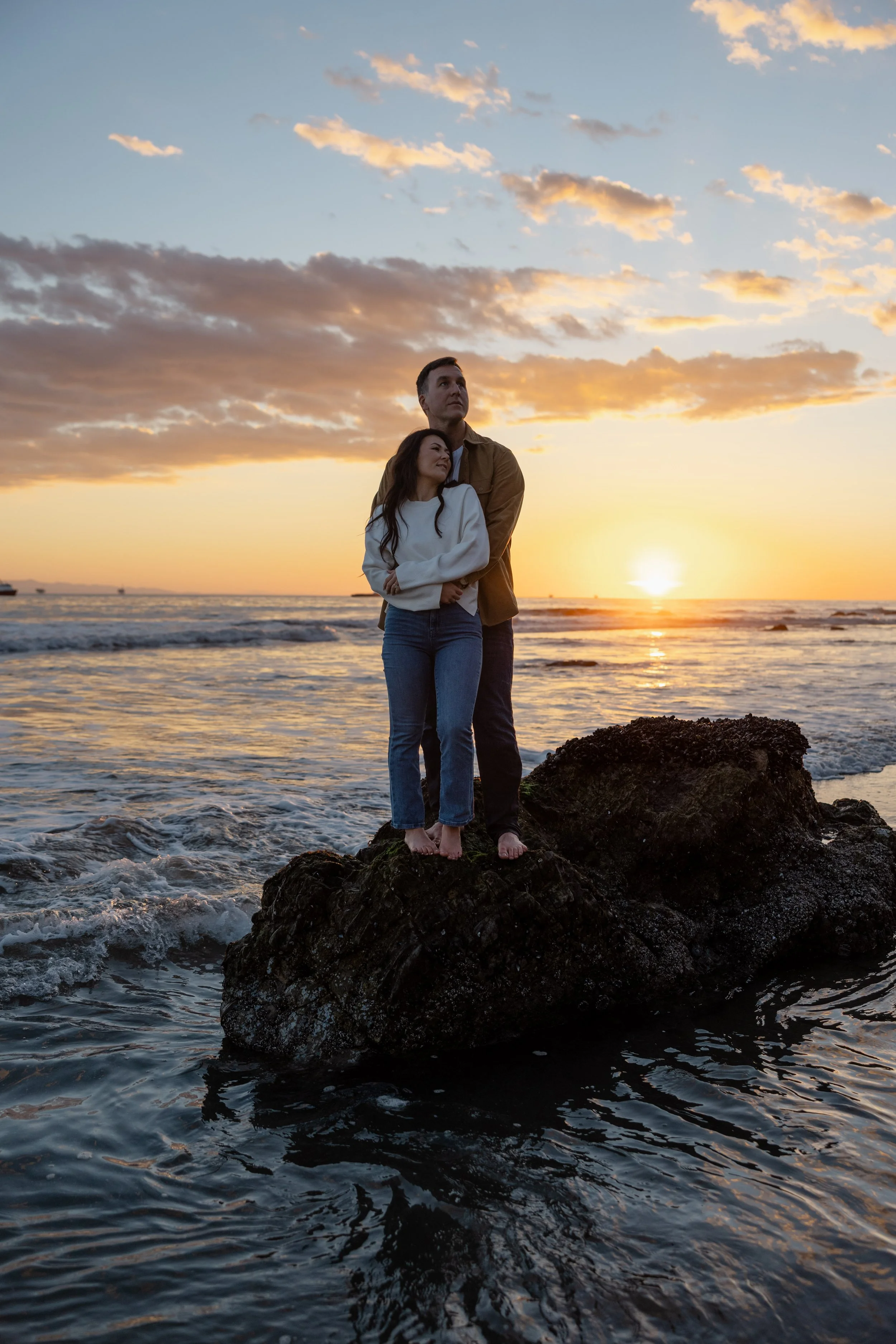 A couple standing on a large rock at the beach during sunset, with the ocean and sky in the background.
