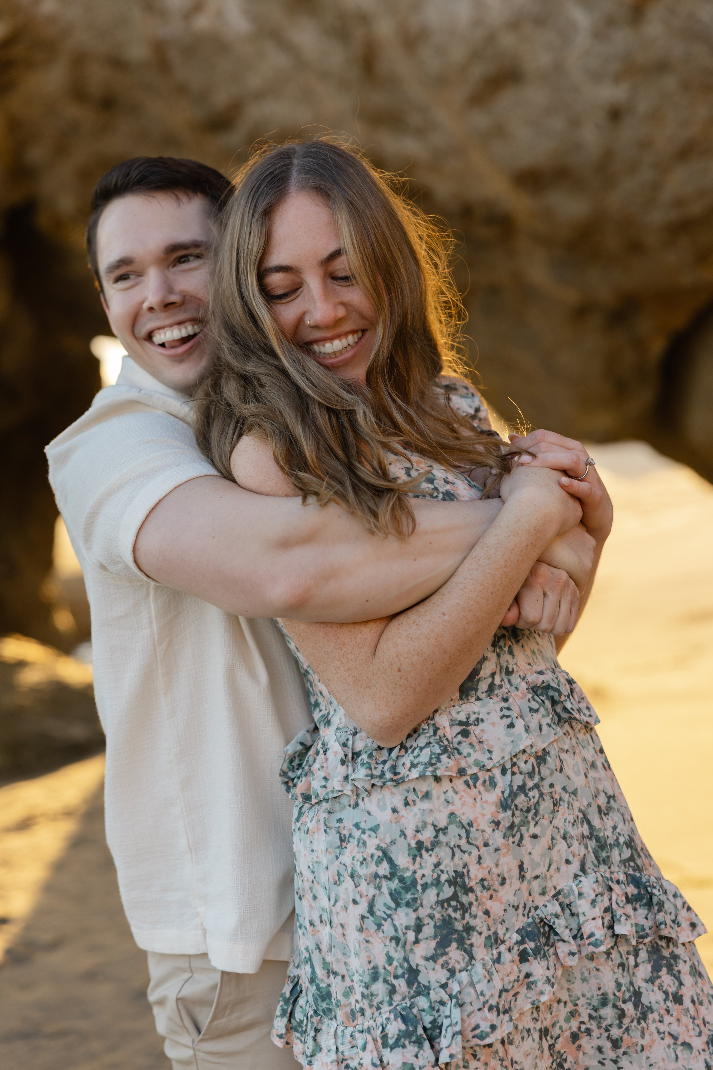 A joyful couple hugging on a beach during sunset, with rocky formations in the background.