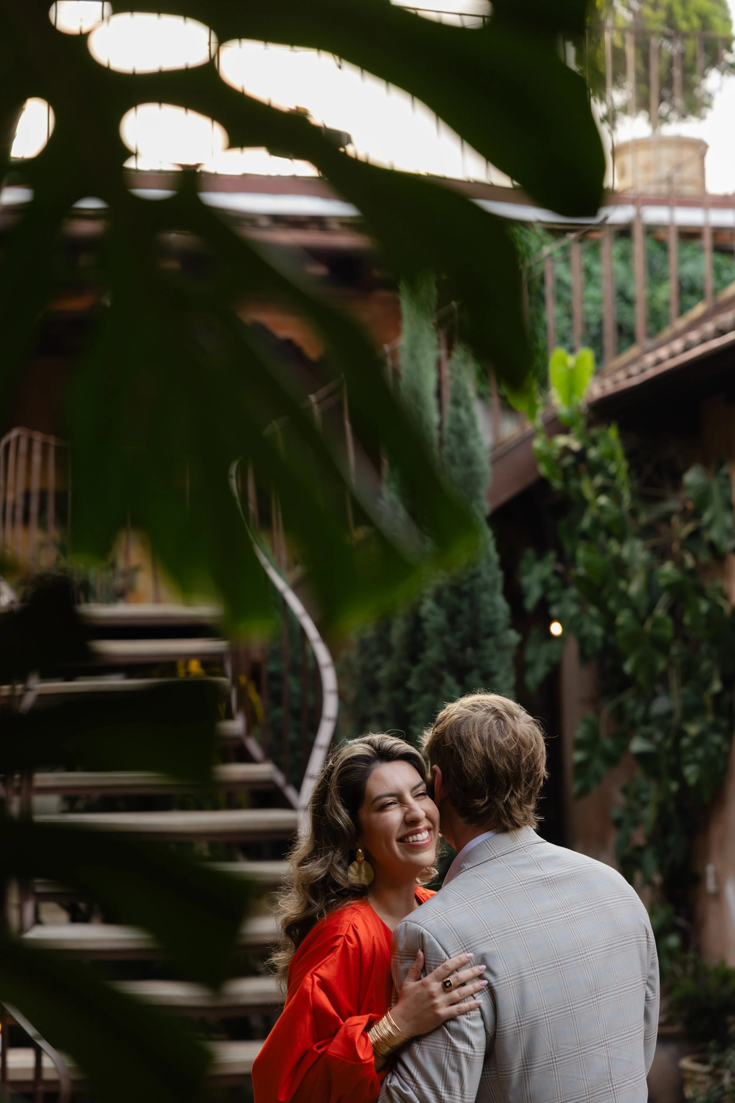 A woman in a red dress smiling and hugging a man in a checkered gray suit in an outdoor garden setting.