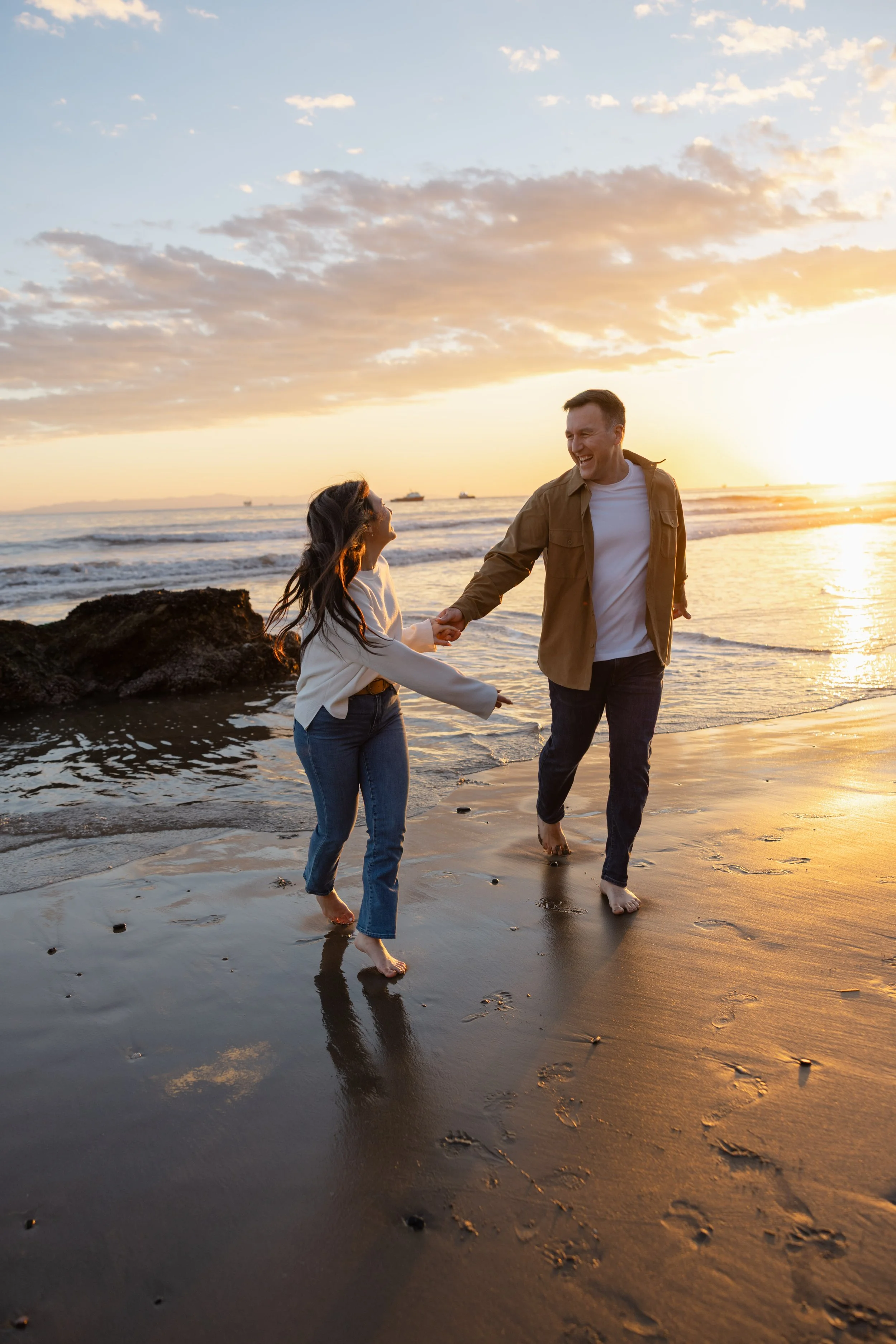 A couple holding hands and smiling while walking on the beach at sunset.