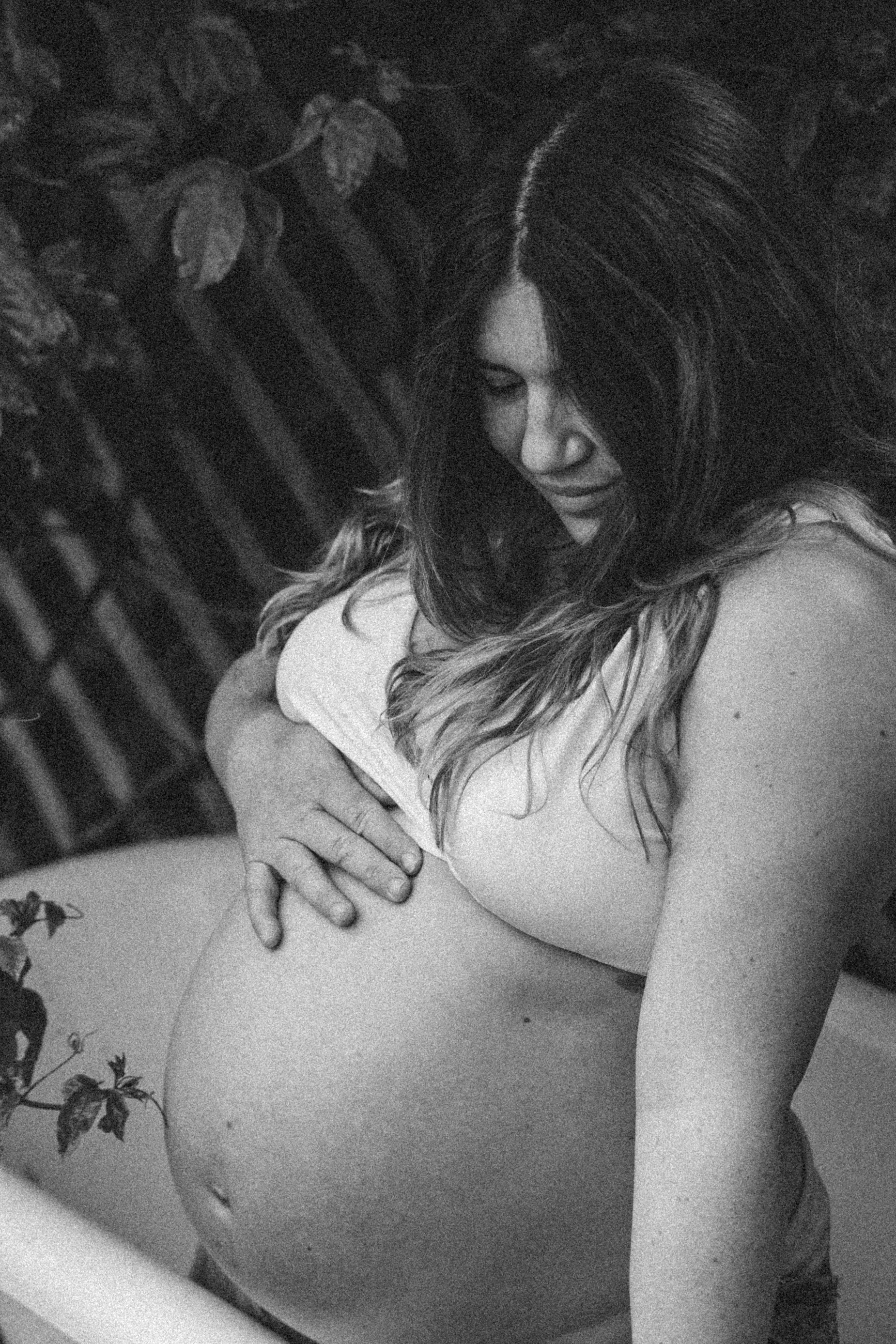 A black-and-white photo of a pregnant woman with long hair, gently cradling her belly in an outdoor setting with foliage.