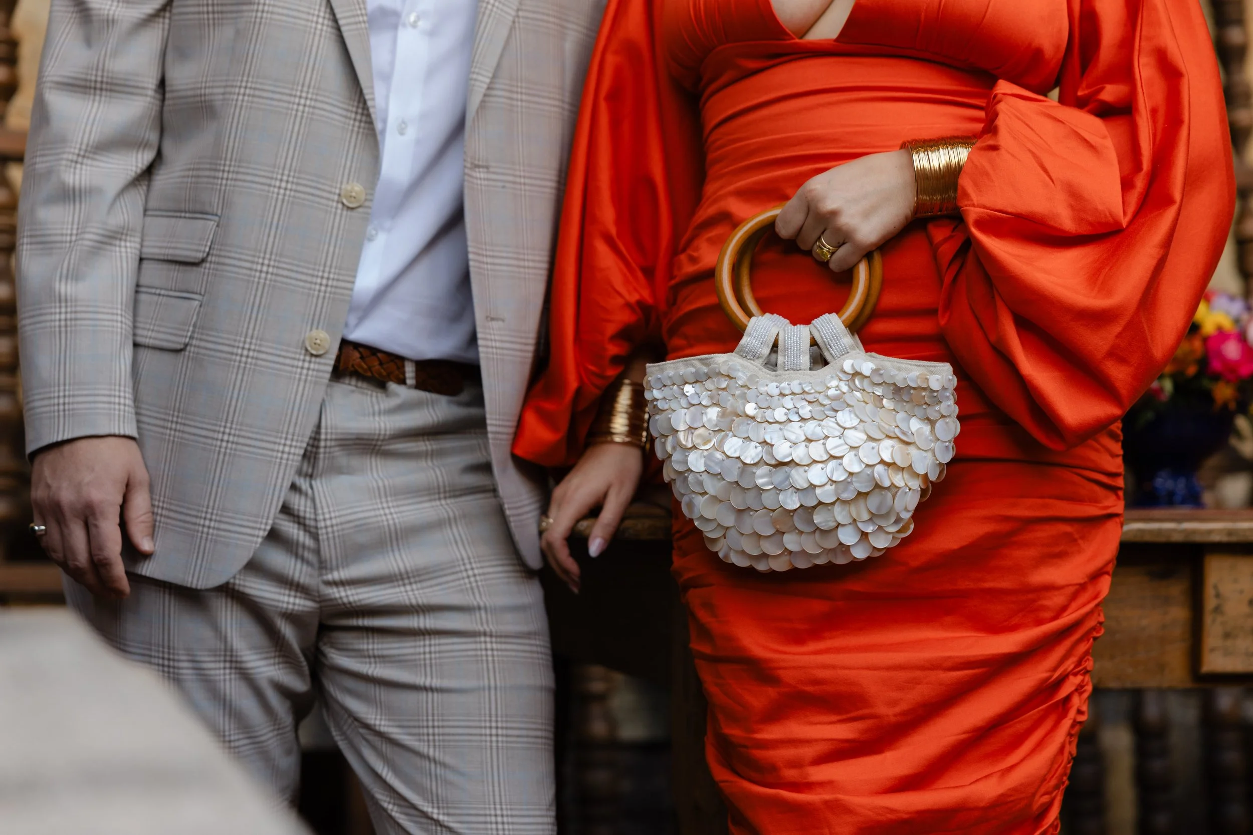 Close-up of a woman in a bright orange dress holding a handbag with white shell sequins and wooden handles, standing next to a man in a light plaid suit.