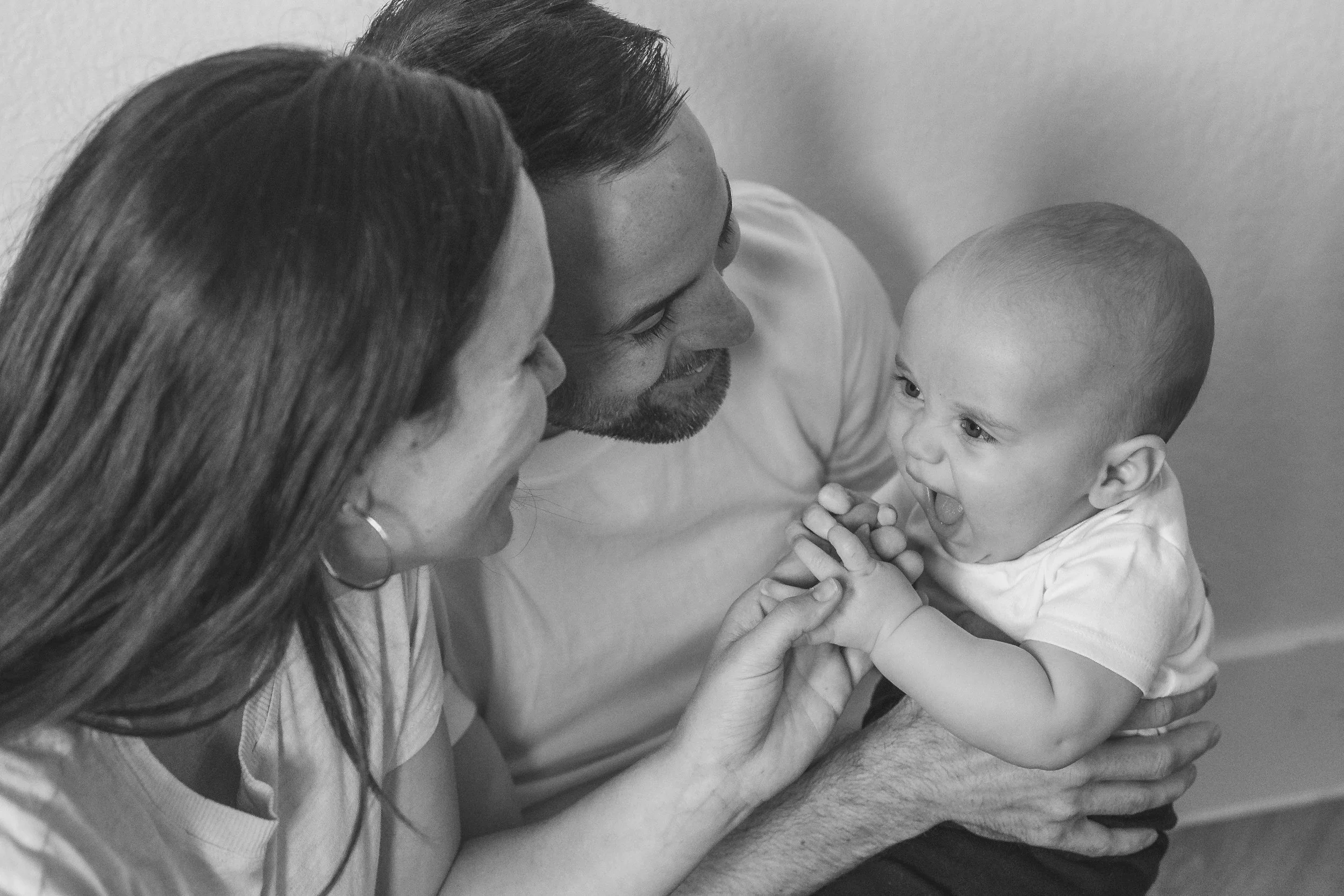 A black and white photo of a family interacting with their baby; a woman, man, and baby are close together, with the woman and man showing affection towards the baby, who appears to be laughing or talking with her mouth open.