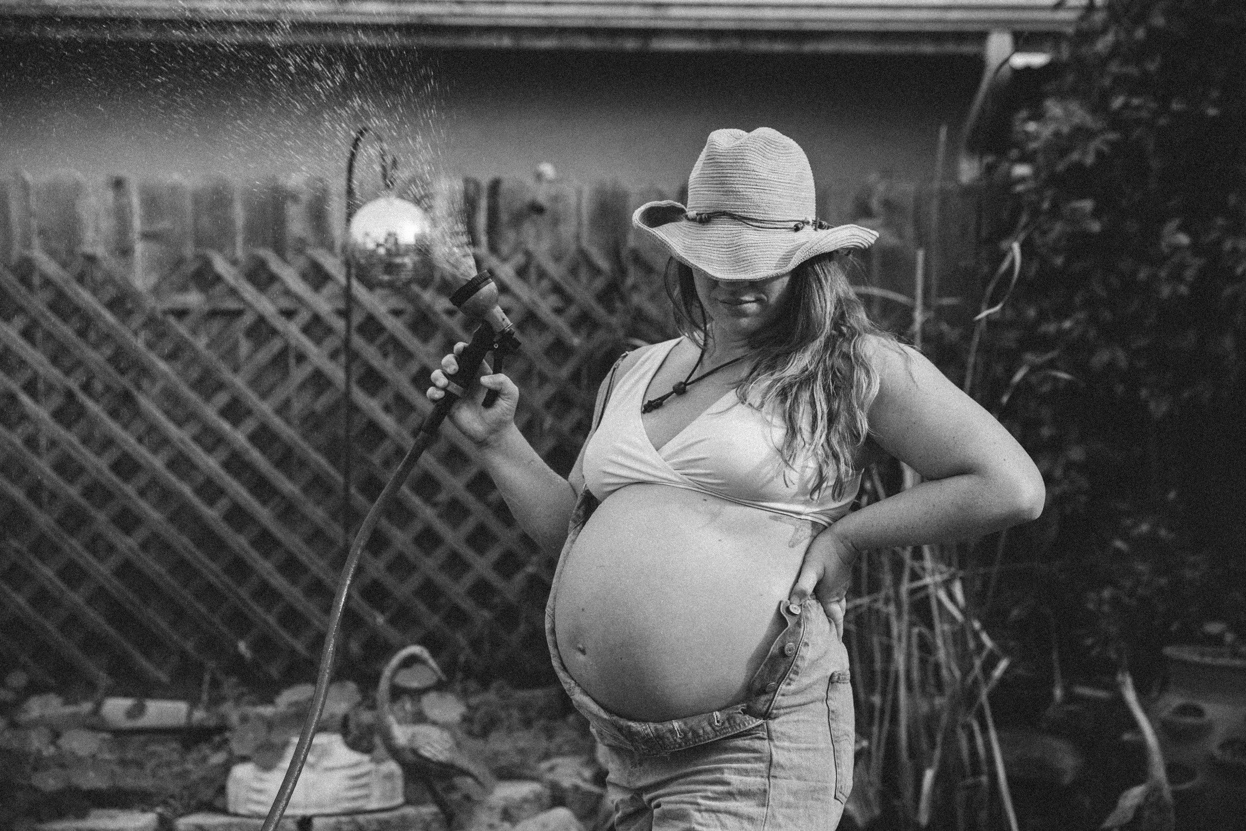 Pregnant woman watering plants in a garden, wearing a wide-brimmed hat and casual clothes, black and white photo.
