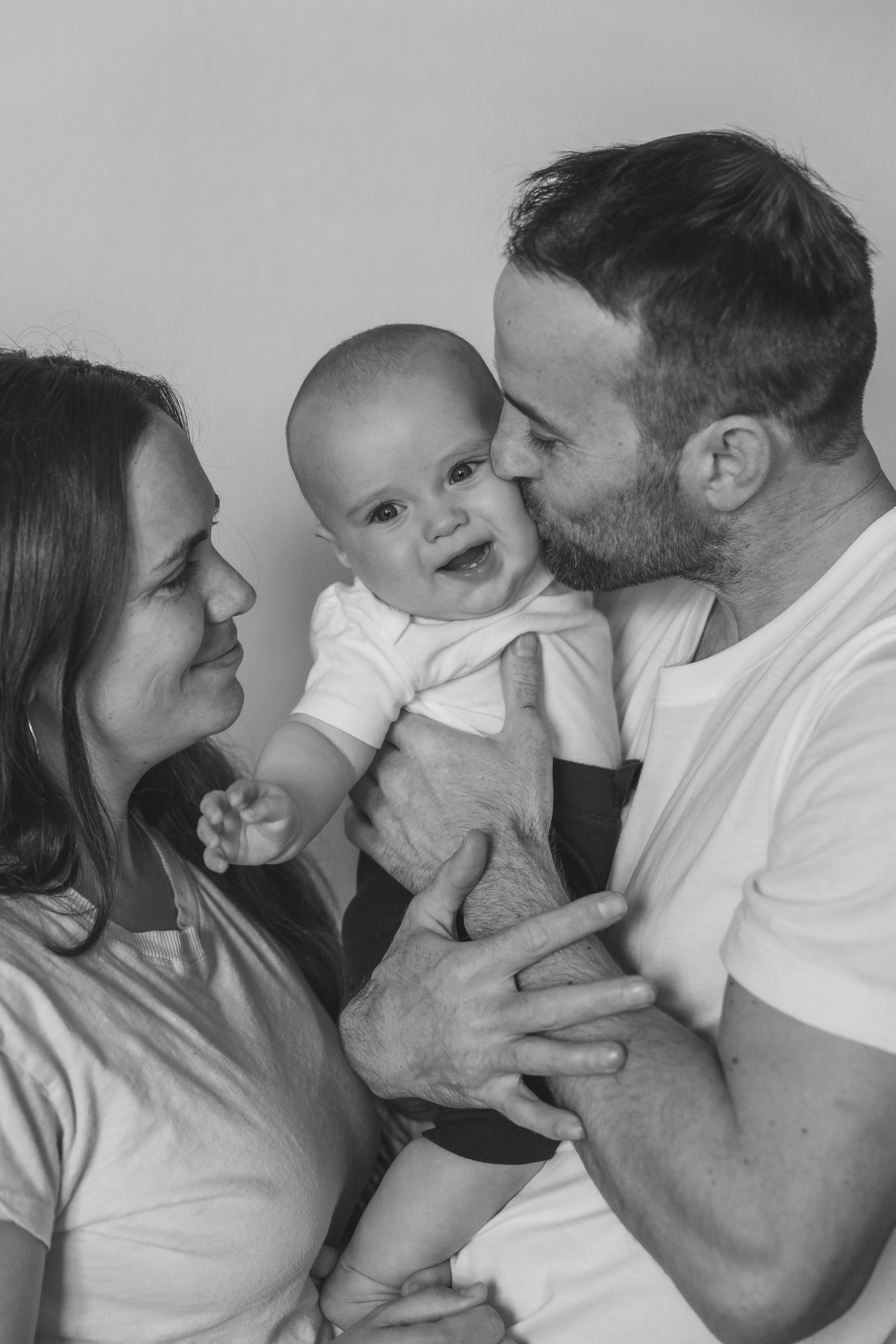A family of three with a baby, the father is kissing the baby on the cheek, the mother looks on smiling. The scene is in black and white.