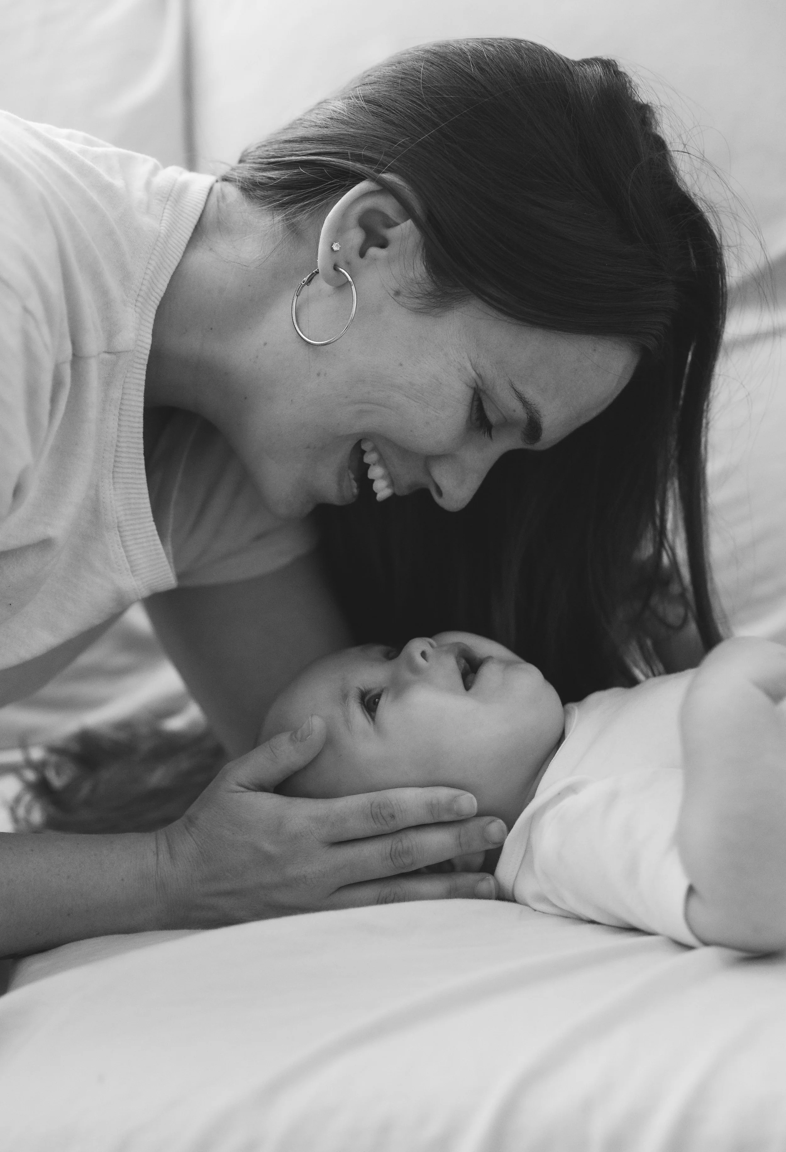 A woman with earrings laughing and leaning over a young child lying on a bed, holding their head gently in a black-and-white photograph.