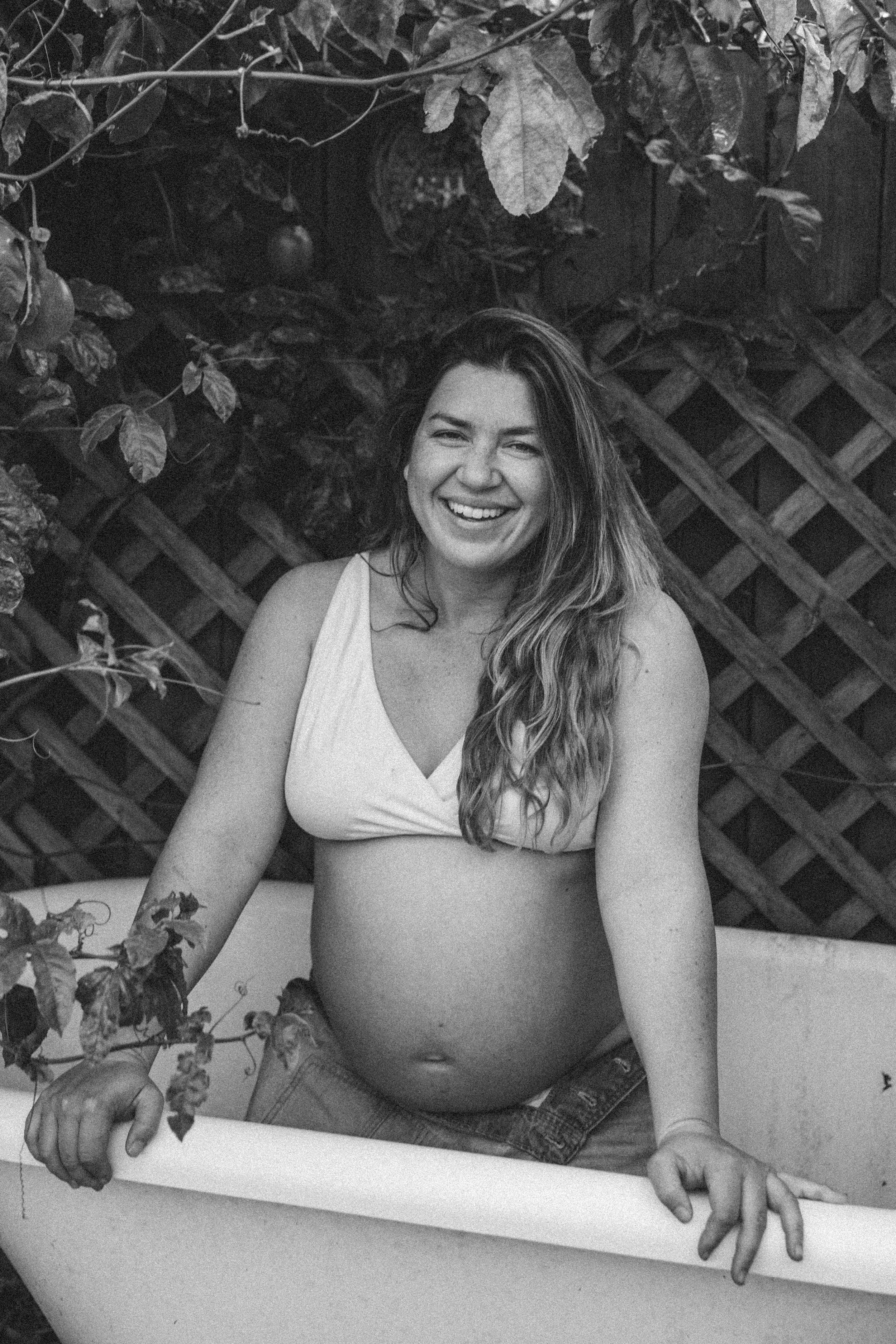 A smiling pregnant woman with long wavy hair sitting in a bathtub outdoors, surrounded by leaves and a wooden lattice in the background.
