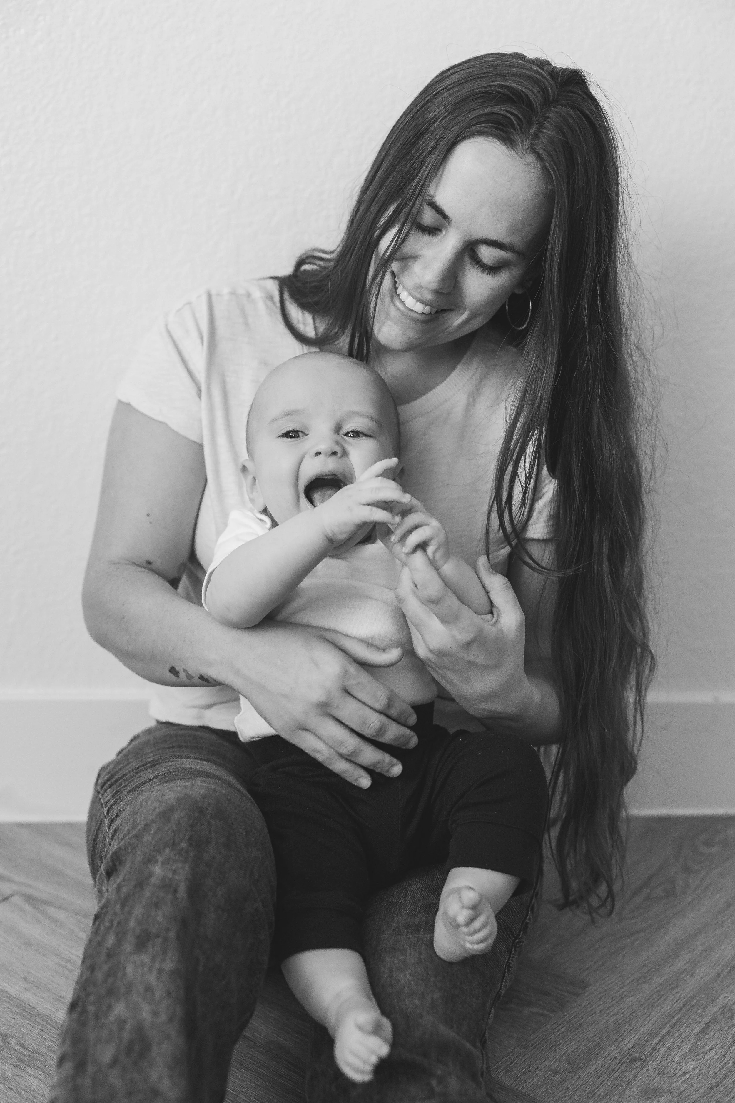 A woman with long hair sitting against a wall, holding a laughing baby on her lap. The woman is smiling and looking down at the baby, who appears to be crying or fussing.
