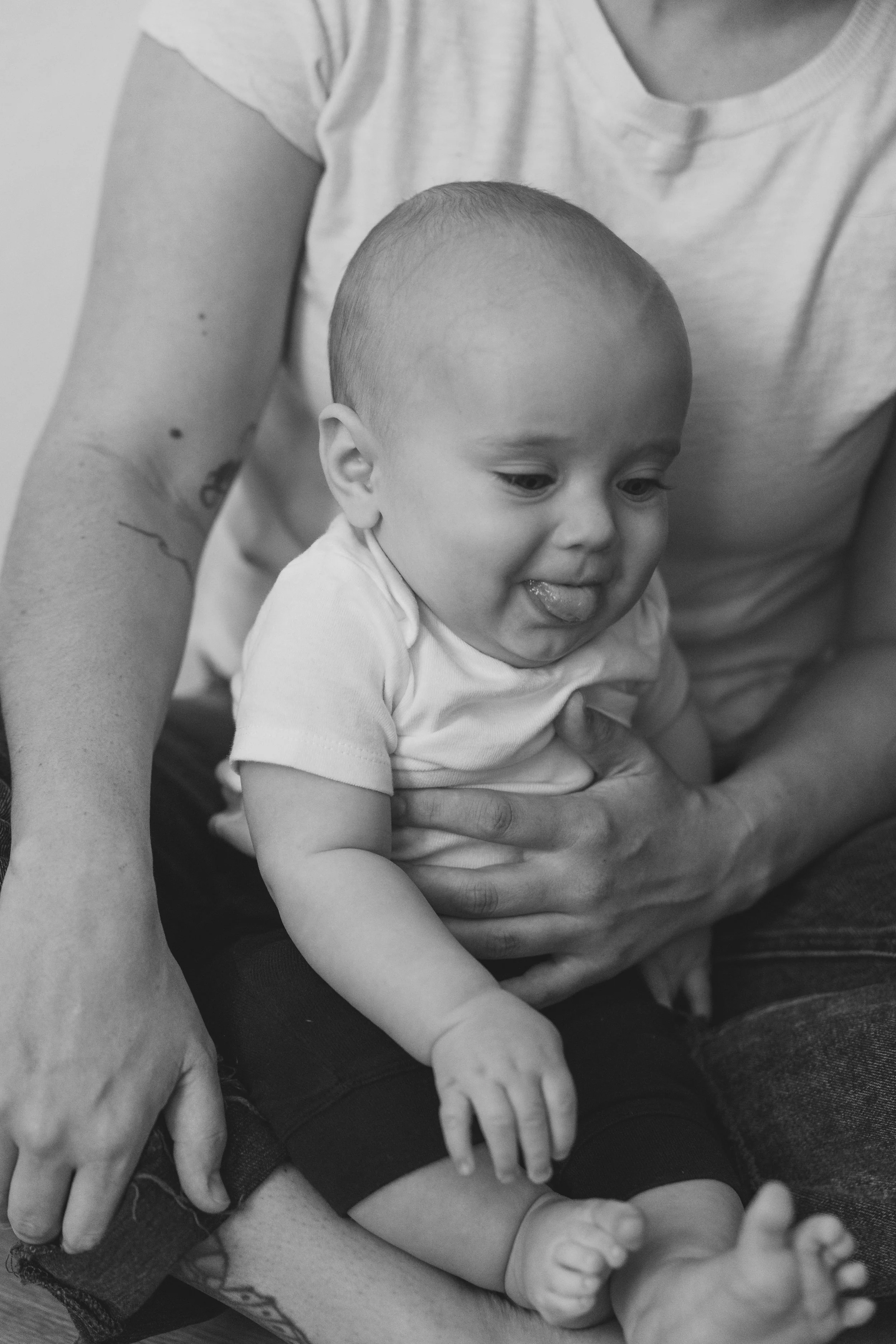 A baby sitting on a person's lap, smiling and sticking out his tongue, in black and white.